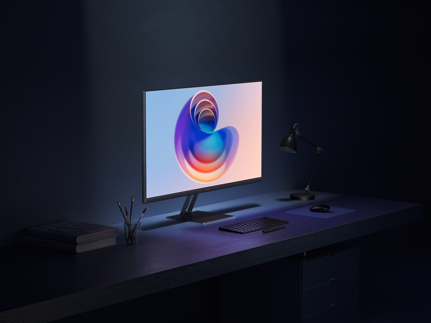 Un monitor de computadora muestra un remolino colorido en un escritorio con teclado, ratón y lámpara. También se ven libros y bolígrafos.