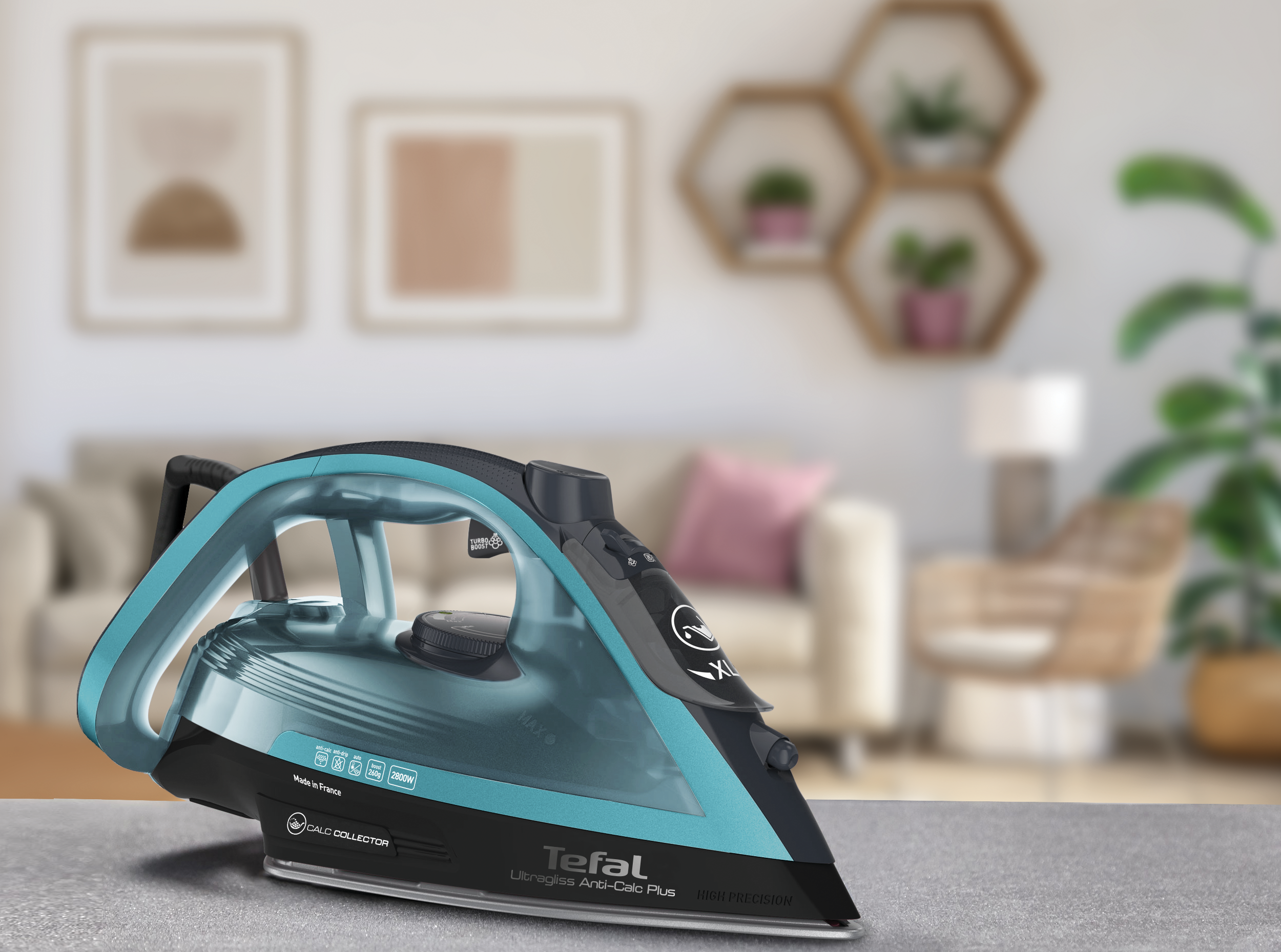 Egy türkizkék és fekete Tefal vasaló egy asztalon. A háttérben egy nappali látható dekorációval. A vasaló van a fókuszban.