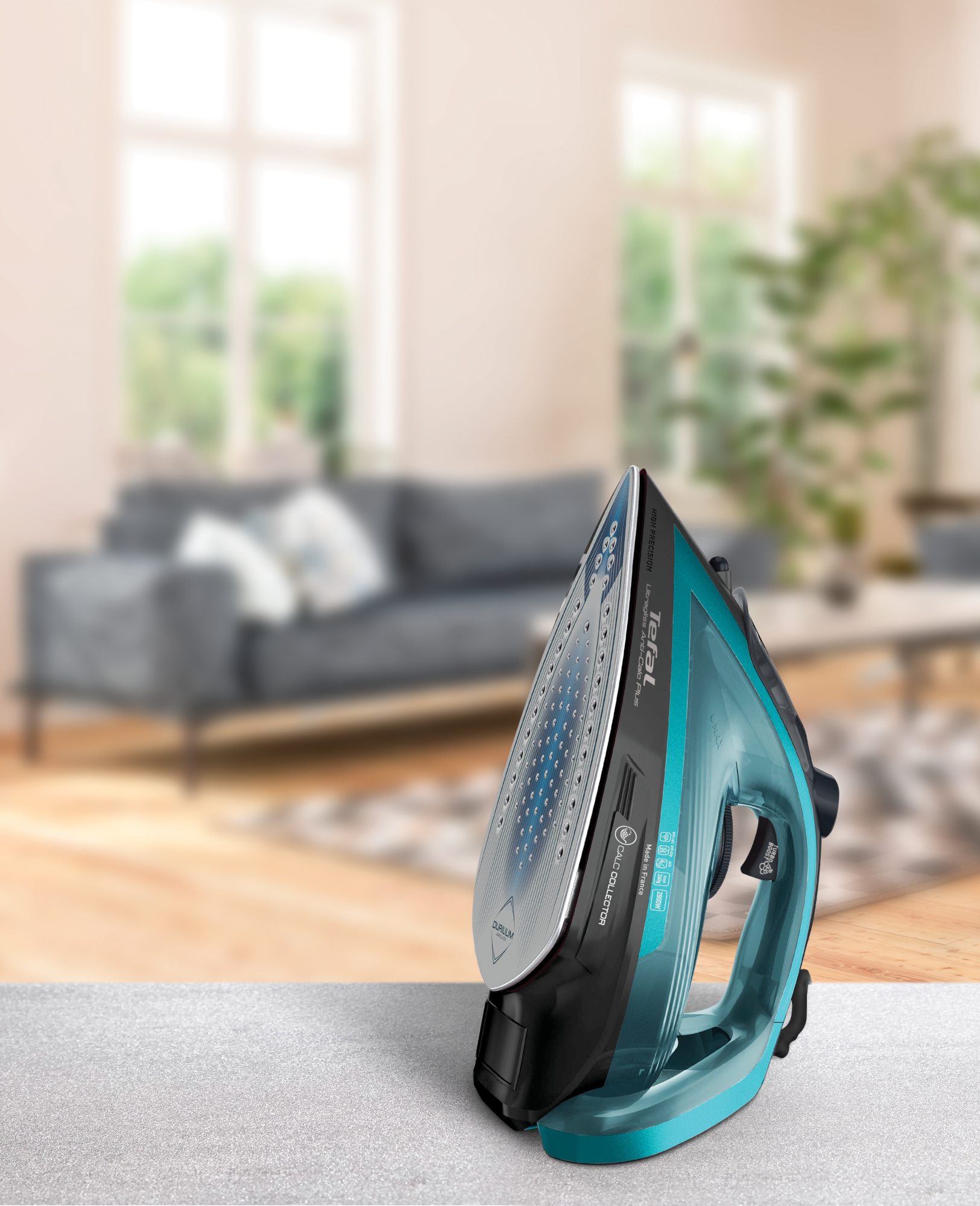 Egy türkizkék és fekete Tefal vasaló szürke felületen. A háttérben egy elmosódott nappali, kanapéval, ablakkal.