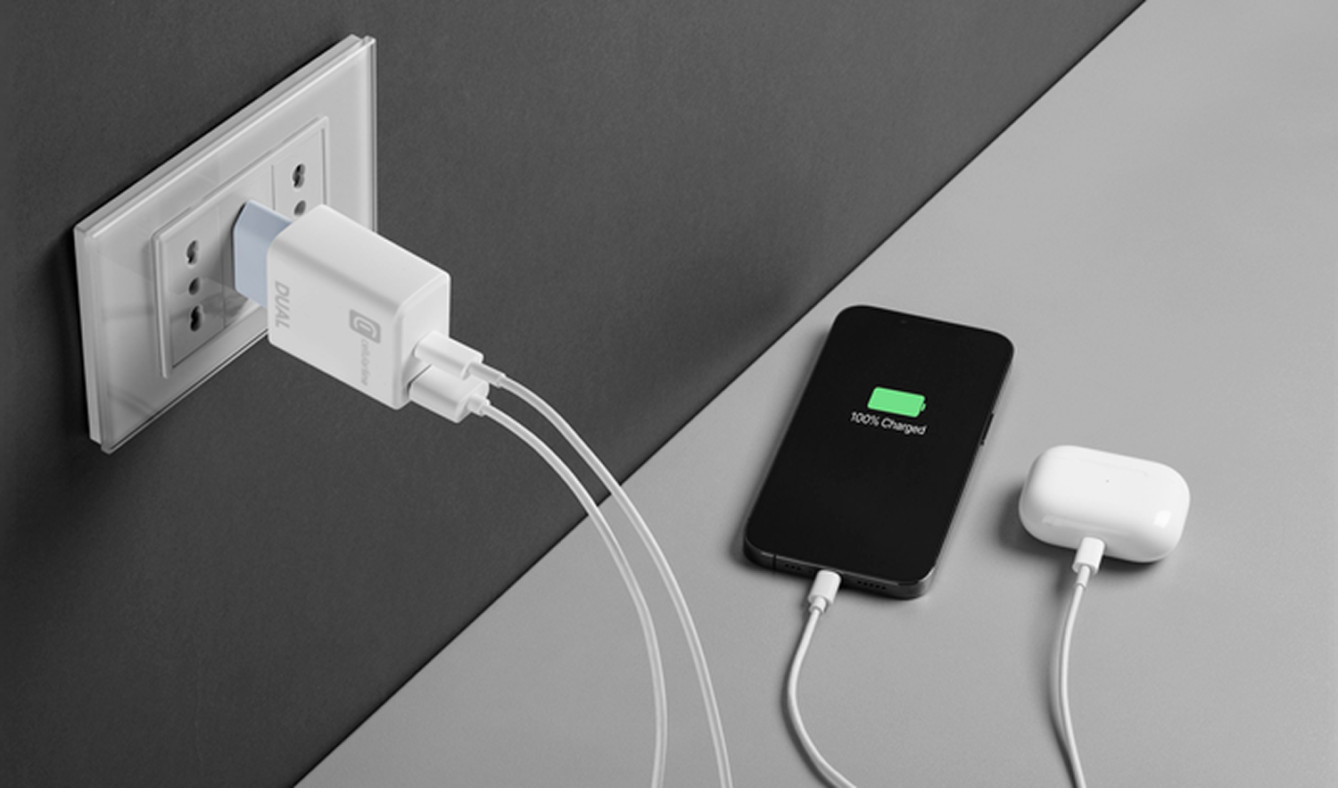 Un cargador USB doble enchufado a una toma de corriente, cargando un teléfono y AirPods. El teléfono muestra 100% cargado.