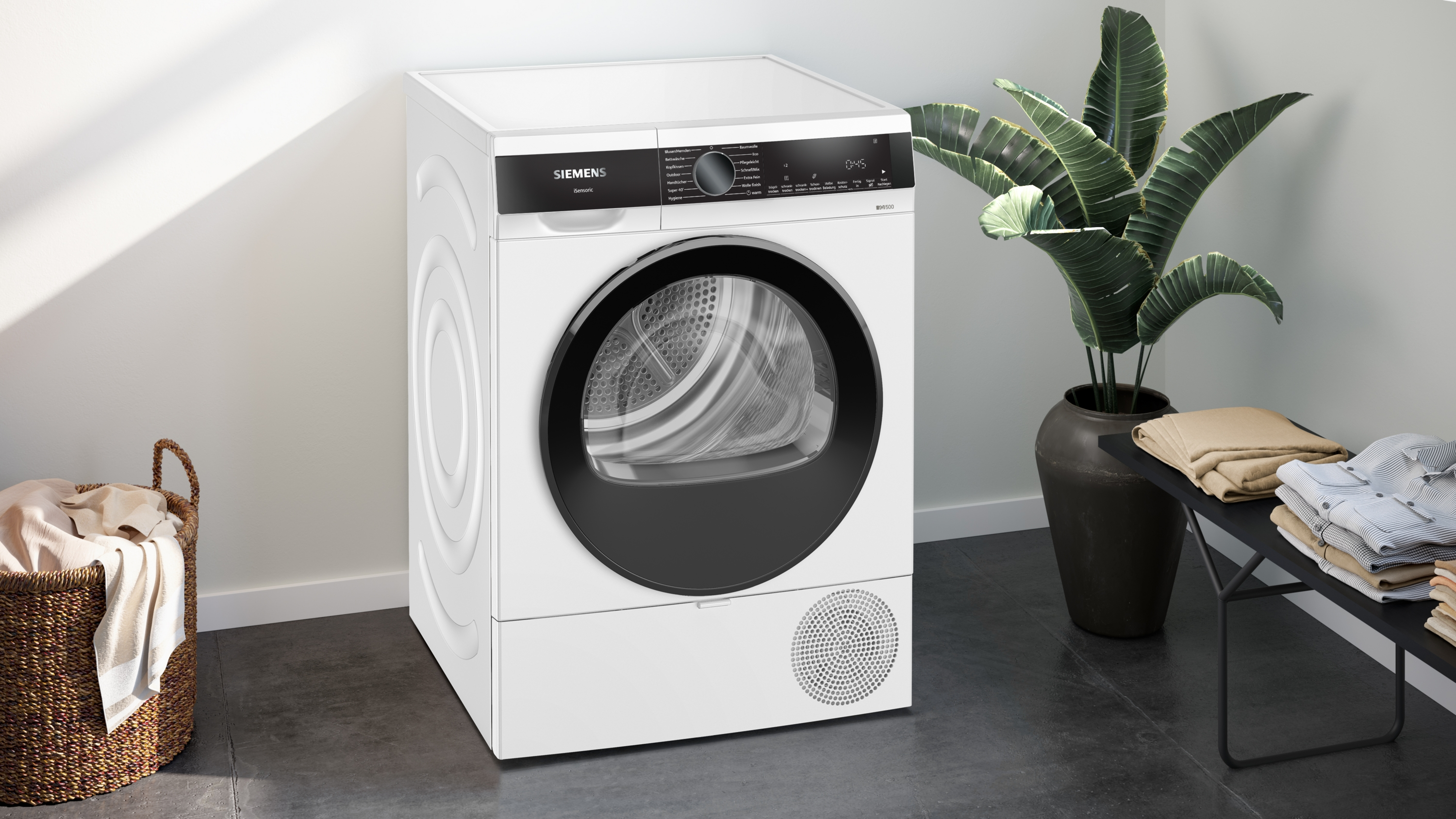 Ein weißer Siemens-Trockner, eine Pflanze, ein Wäschekorb und ein schwarzer Tisch mit Kleidung.