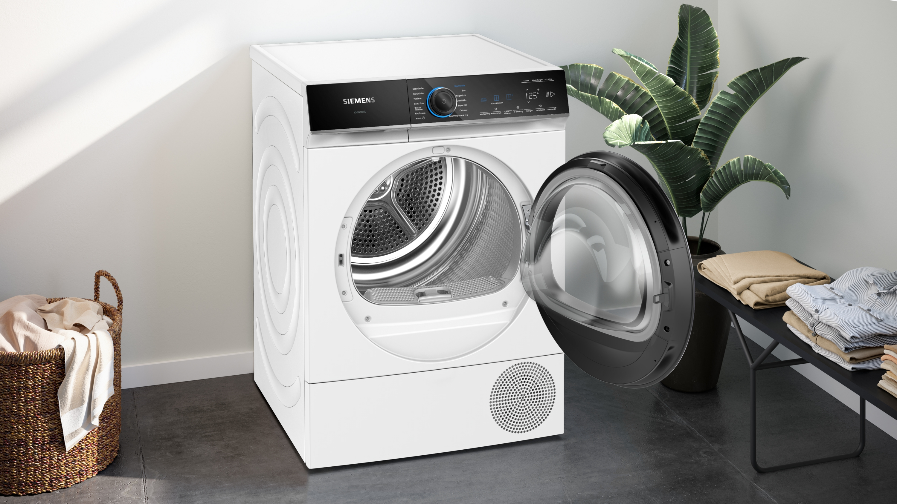 Ein weißer Siemens-Trockner mit offener Tür. Daneben eine Pflanze, ein Wäschekorb und gefaltete Kleidung. Der Hintergrund ist eine weiße Wand.