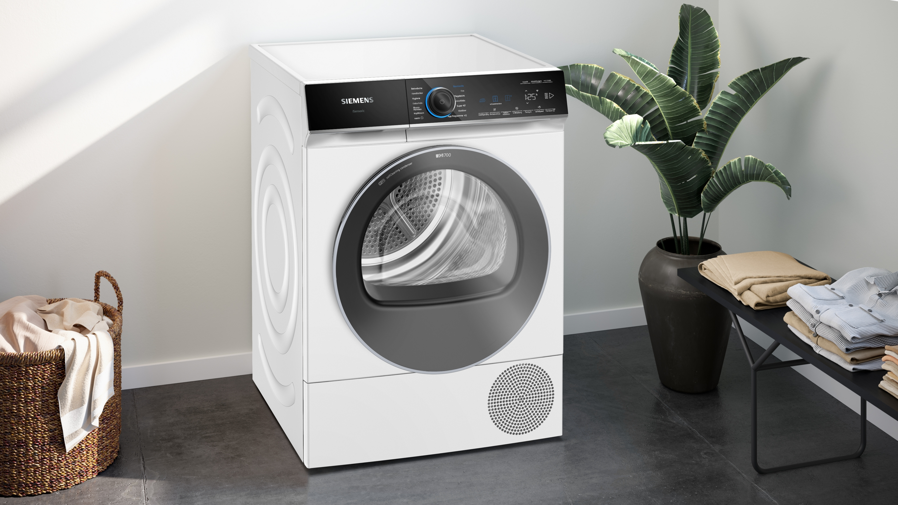 Ein weißer Siemens-Trockner steht neben einer Pflanze und einem Tisch mit gefalteter Kleidung. Links steht ein Wäschekorb.