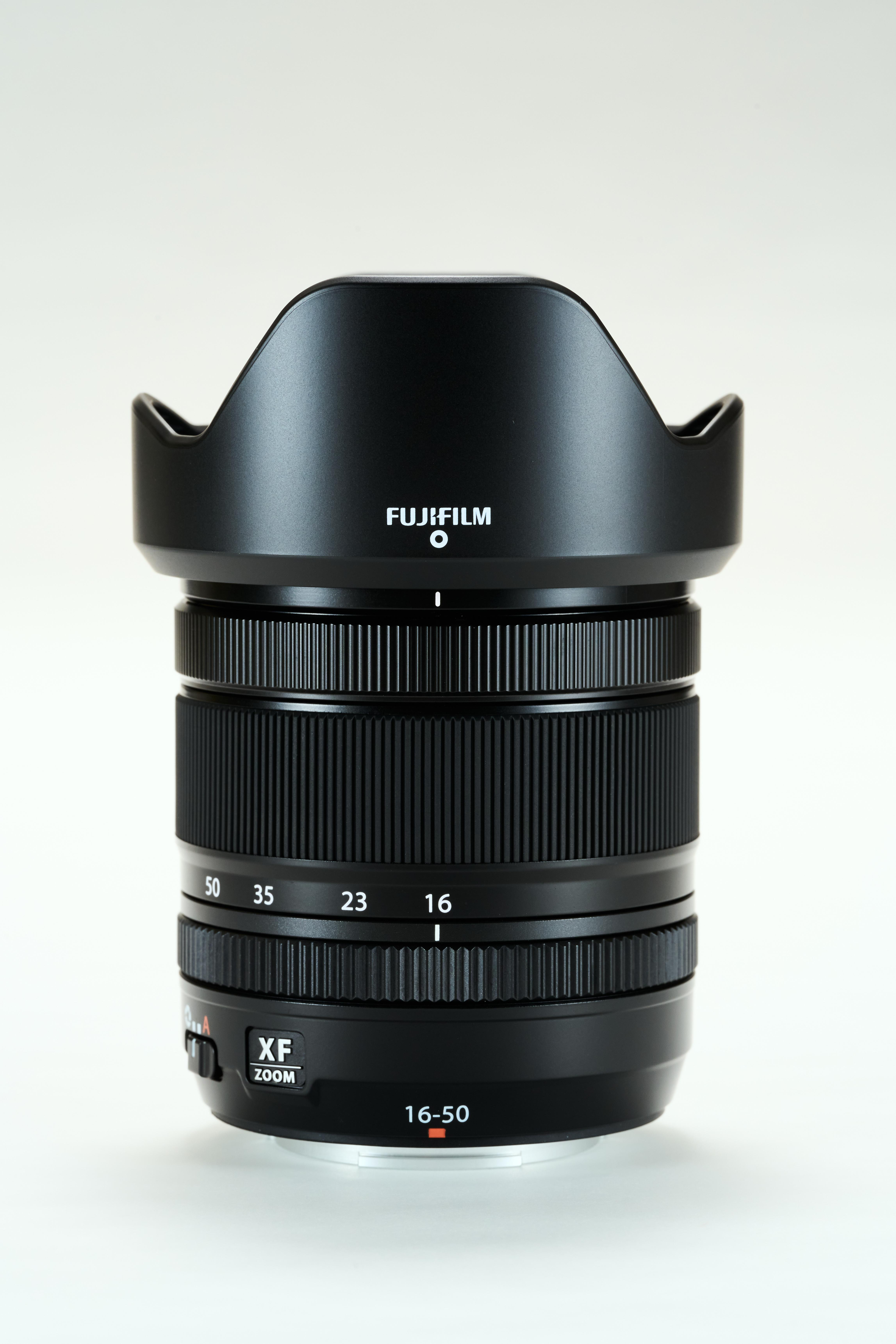 Schwarzes Fujifilm-Kameraobjektiv mit Gegenlichtblende. Es hat Zoomeinstellungen und das Objektiv befindet sich auf weißem Hintergrund.