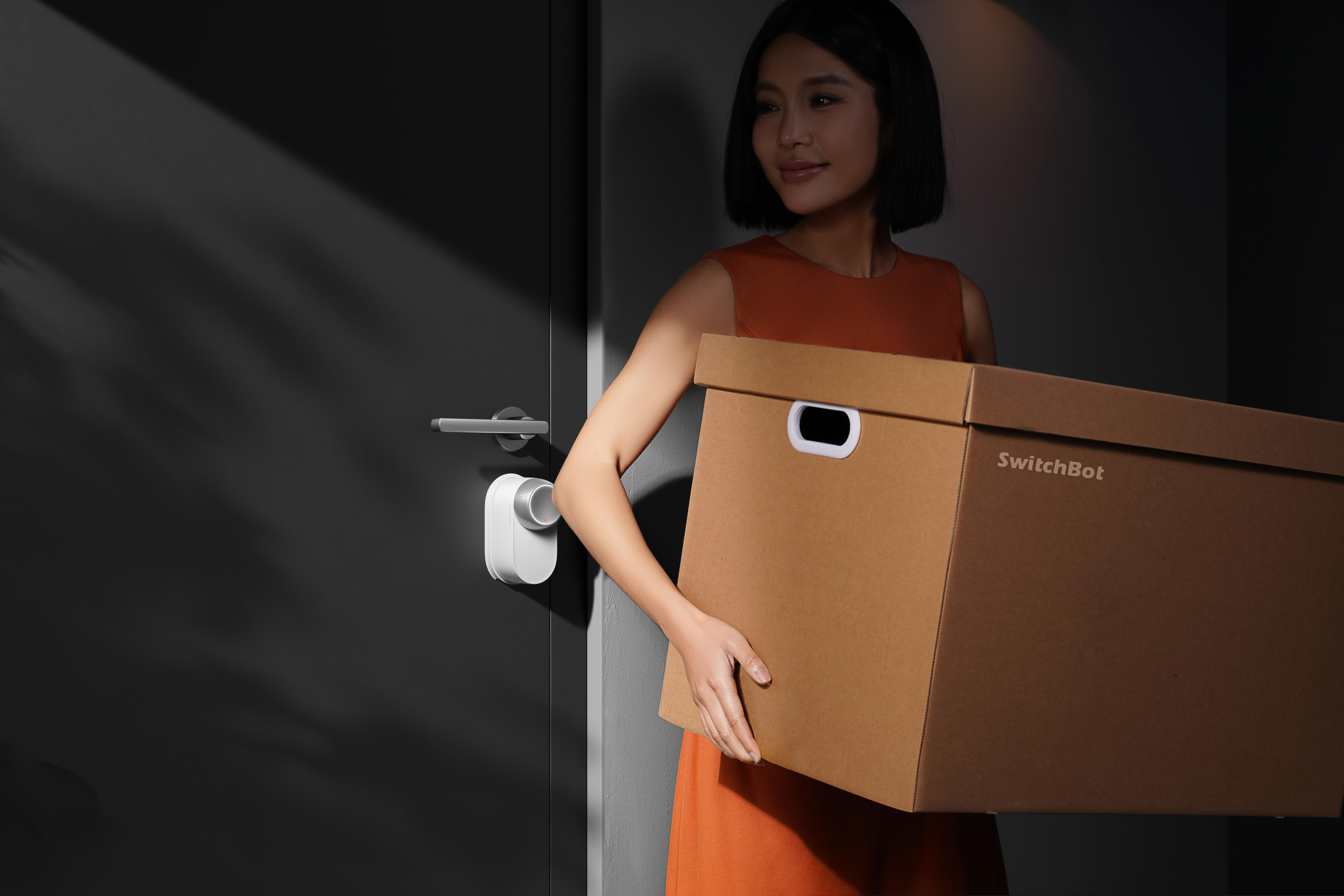 Eine Frau in Orange hält eine SwitchBot-Box in der Nähe einer Tür mit einem intelligenten Schloss. Die Tür ist dunkelgrau.