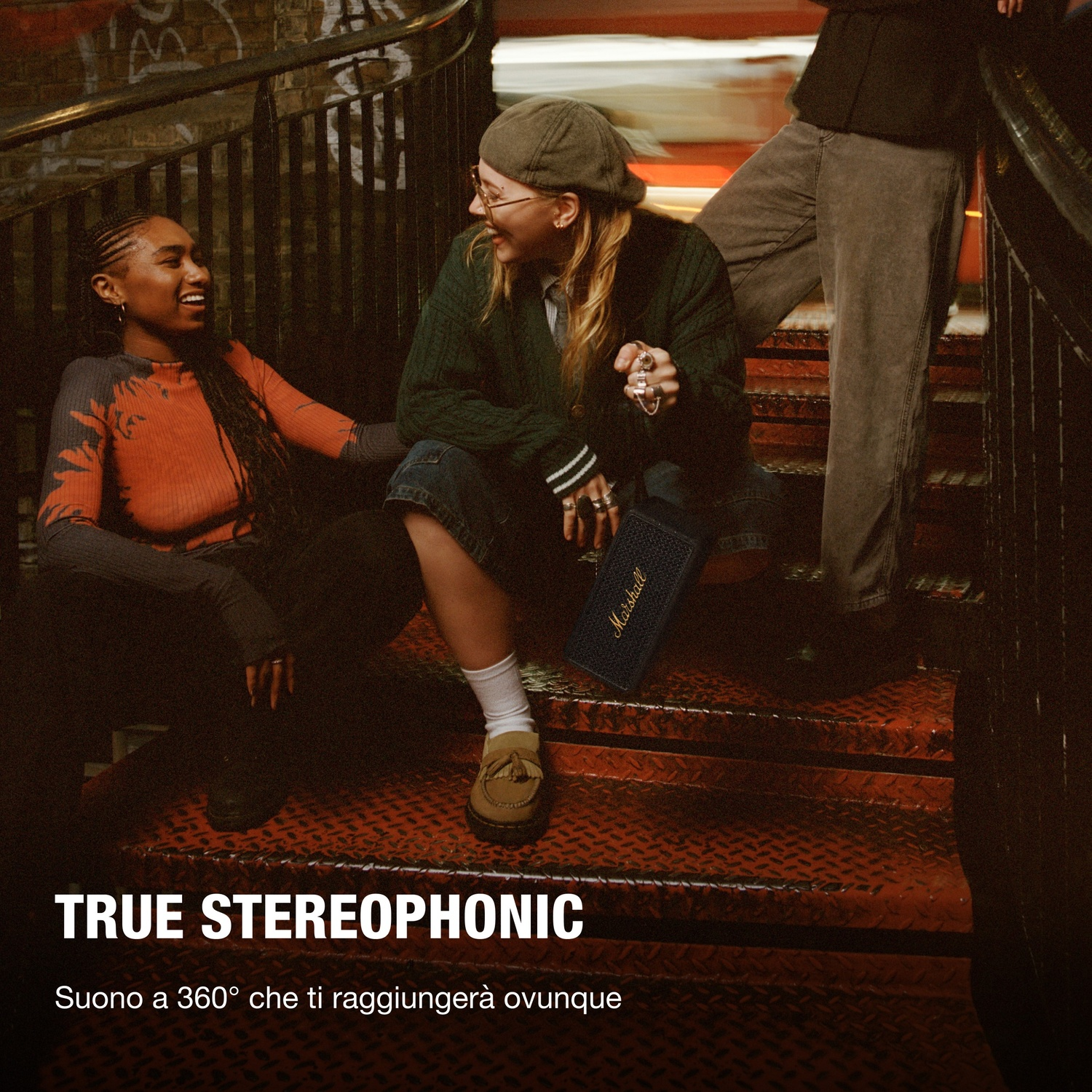 Tre persone su scale metalliche ridono. Una tiene un altoparlante Marshall. Graffiti su muro di mattoni. Testo "True stereophonic" visibile.