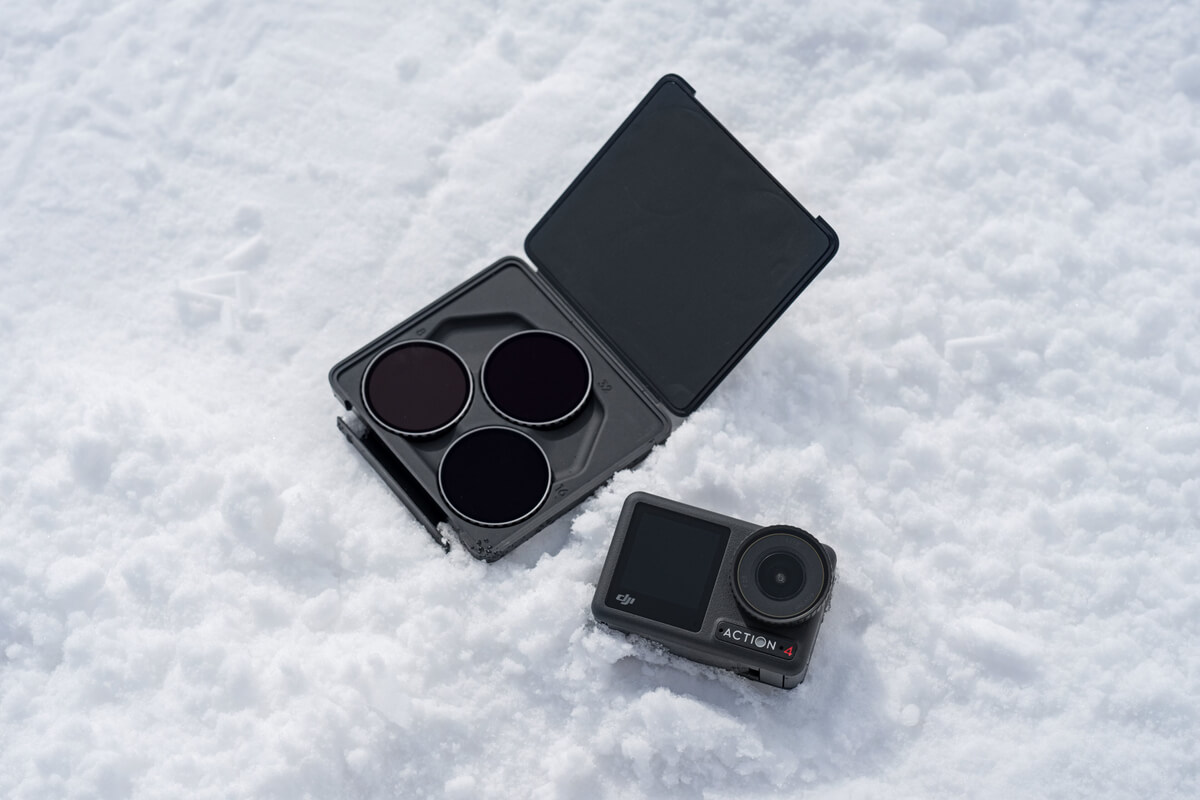 DJI Action 4 kamera ve filtre çantası kar üzerinde. Kamera siyah, filtreler koyu. Arka plan beyaz kar.