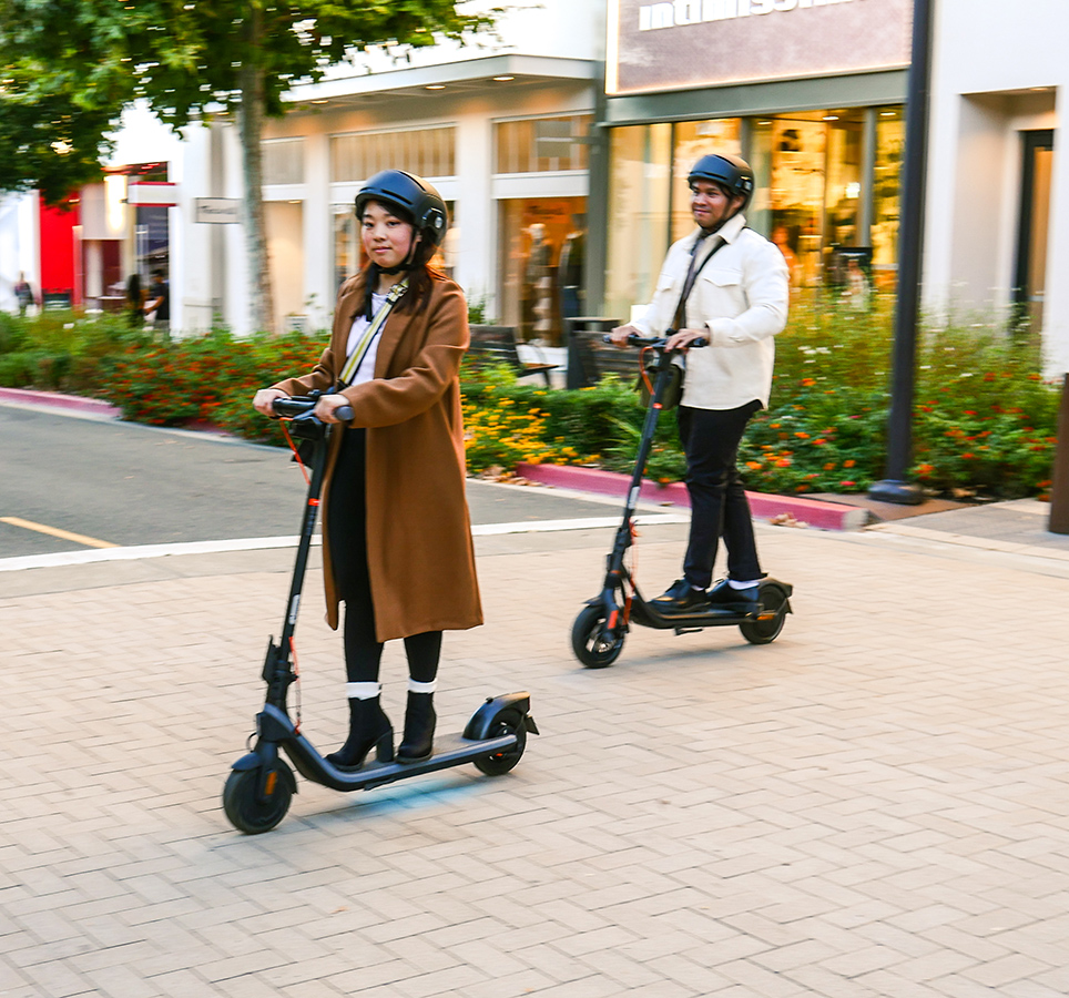 İki kişi, tuğla bir yolda elektrikli scooter kullanıyor. Kask ve mont giyiyorlar. Arka planda dükkanlar ve ağaçlar var.