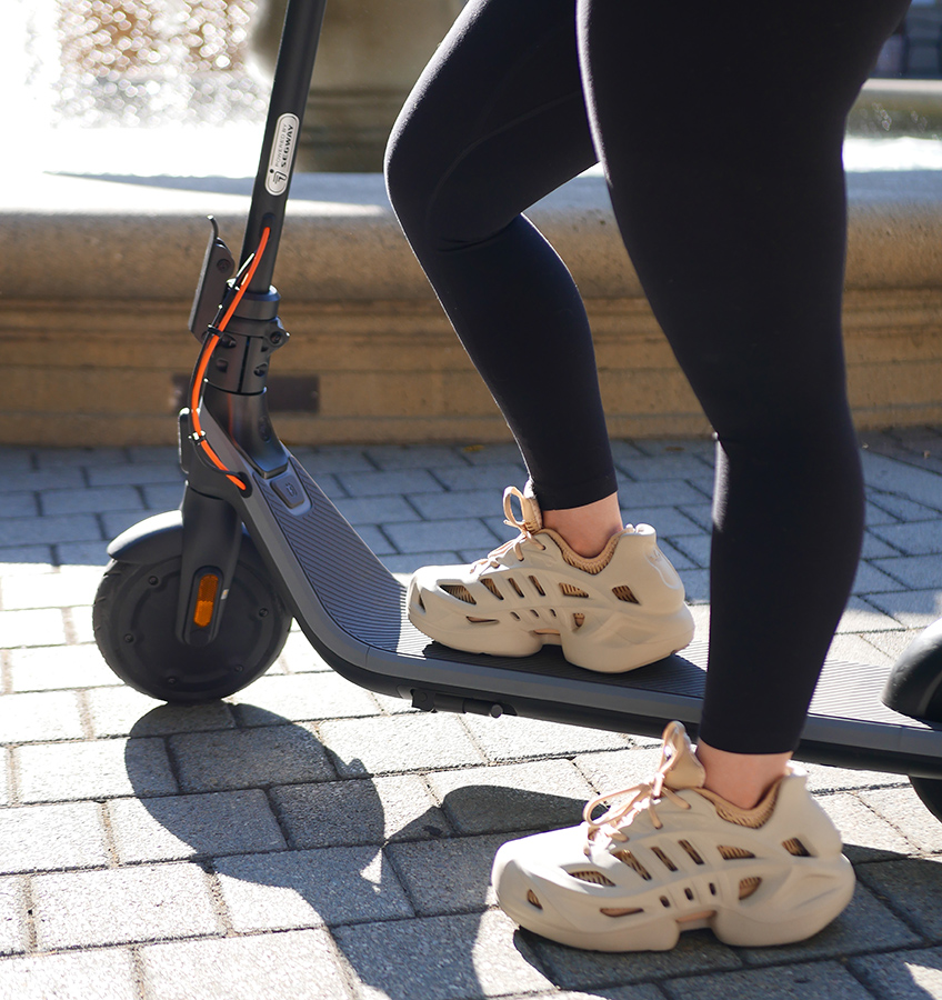 Siyah tayt ve bej spor ayakkabı giyen, scooter kullanan kişi. Scooter, tuğla yüzey üzerinde siyah ve gri renktedir.