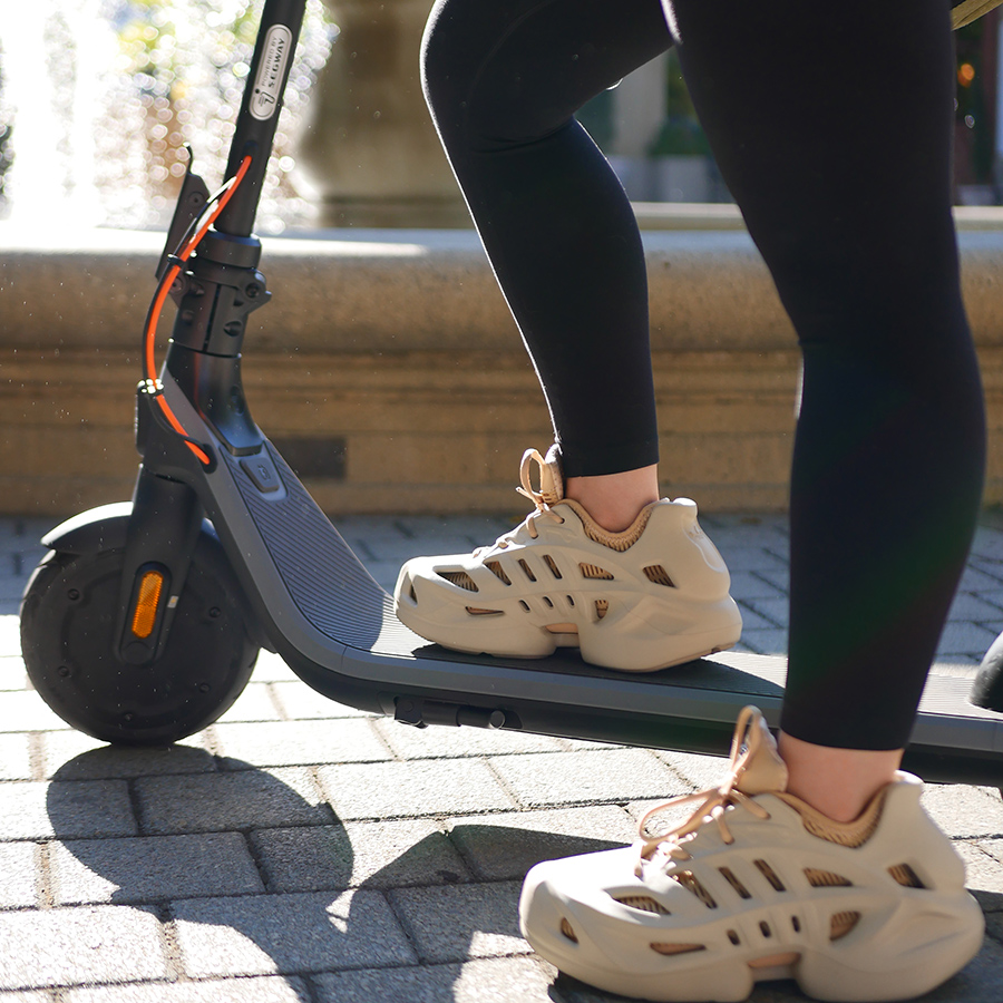 Bir kişi, bej spor ayakkabı ve siyah tayt giyerek, tuğla bir yolda, arka planda bir çeşme ile elektrikli scooter kullanıyor.