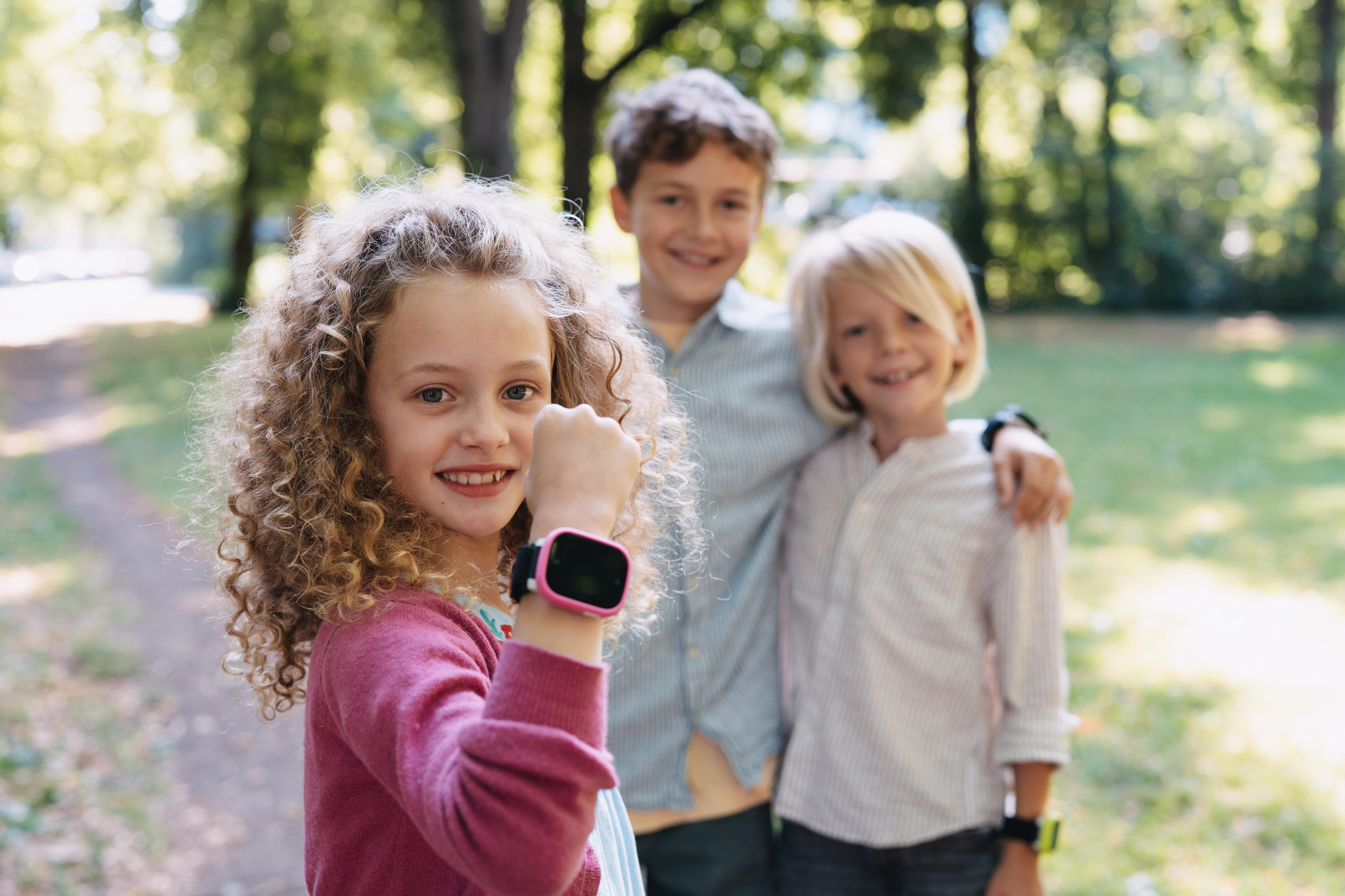 Drei lächelnde Kinder im Park. Ein Mädchen in Rosa zeigt ihre Smartwatch. Zwei Jungen stehen sich umarmend gegenüber.