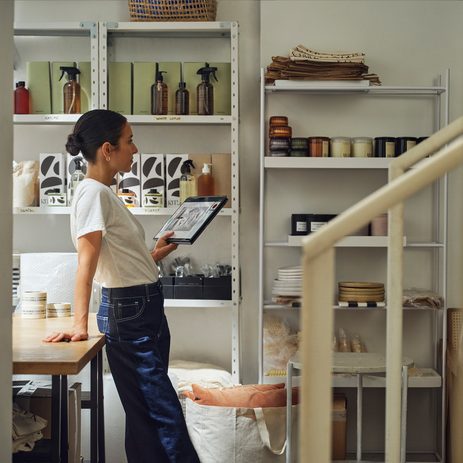 A woman in a white shirt and jeans examines a tablet. Shelves behind her hold products and supplies. Staircase and white walls are visible.