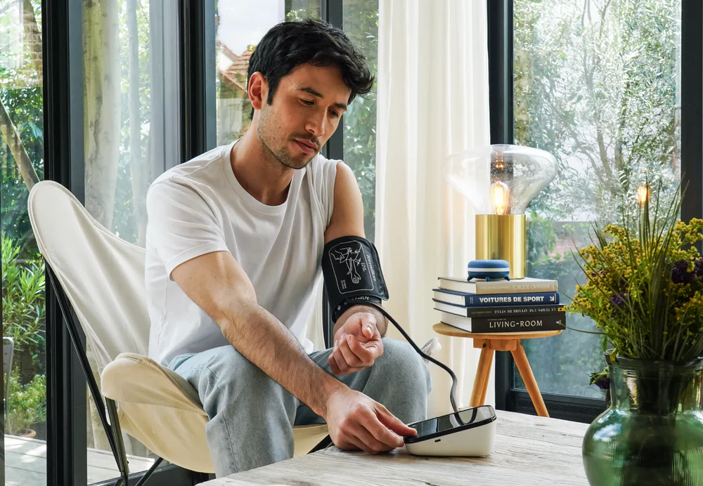 Un homme en t-shirt blanc prend sa tension artérielle. Il est dans une pièce moderne avec une chaise, une lampe, des livres et des fleurs.