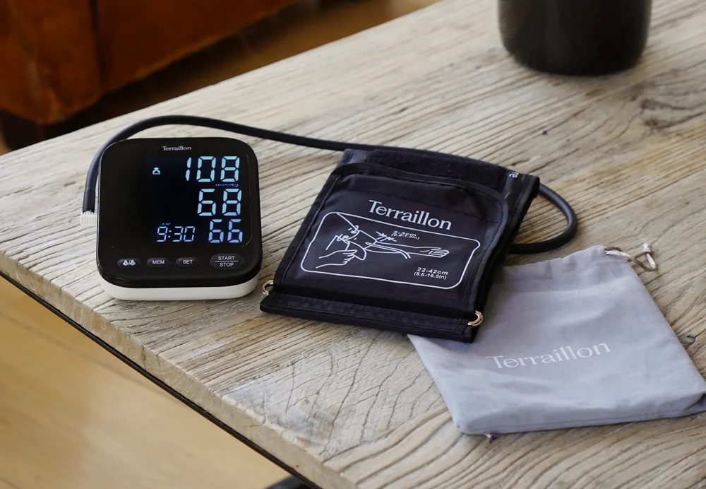 Tensiomètre avec brassard et pochette sur une table en bois. L'écran affiche les lectures. Comprend un brassard noir et une pochette grise.