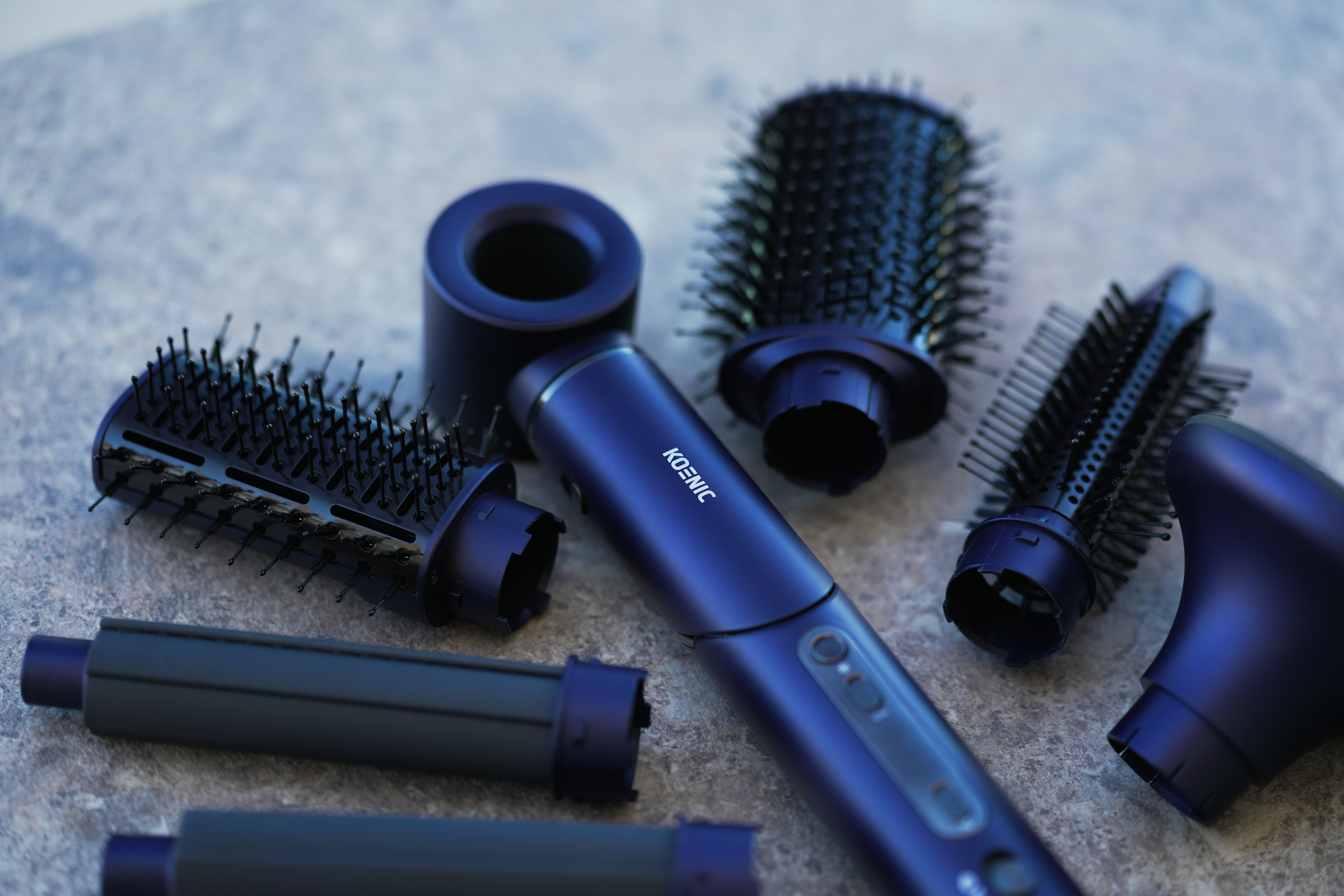 Un styler capillaire Koenic avec plusieurs accessoires. Les accessoires bleus et noirs sont posés sur une surface grise. Le styler est également bleu.