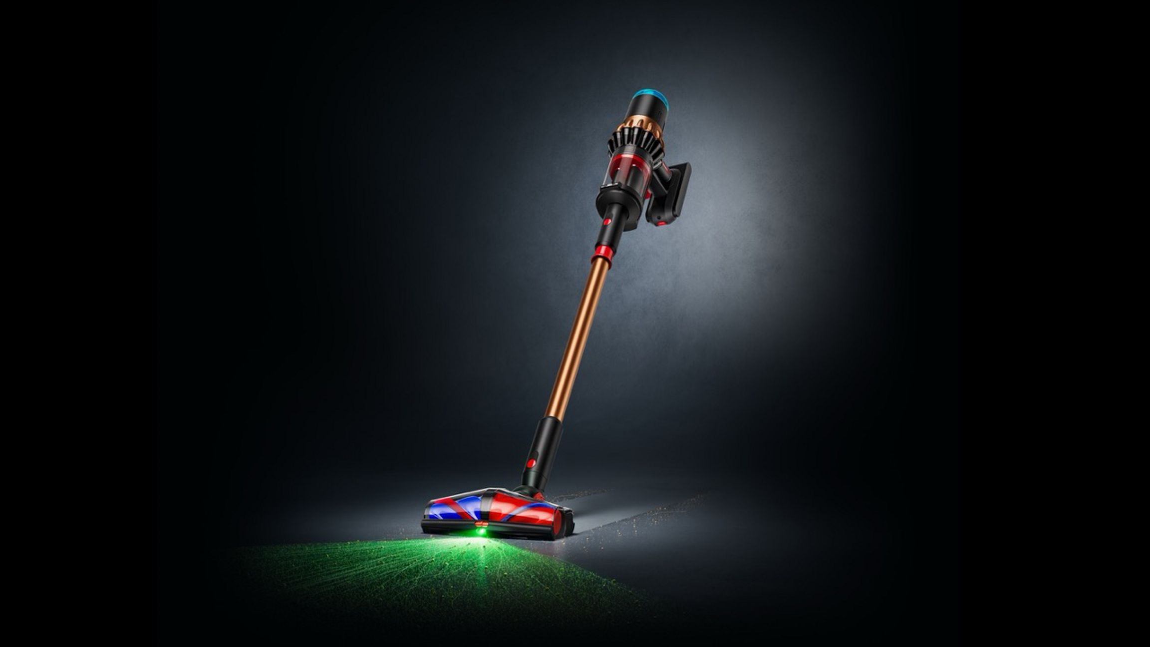Un aspirateur sans fil est posé sur une surface sombre. Il a une baguette en bronze, une poignée noire et une tête de nettoyage rouge et bleue.