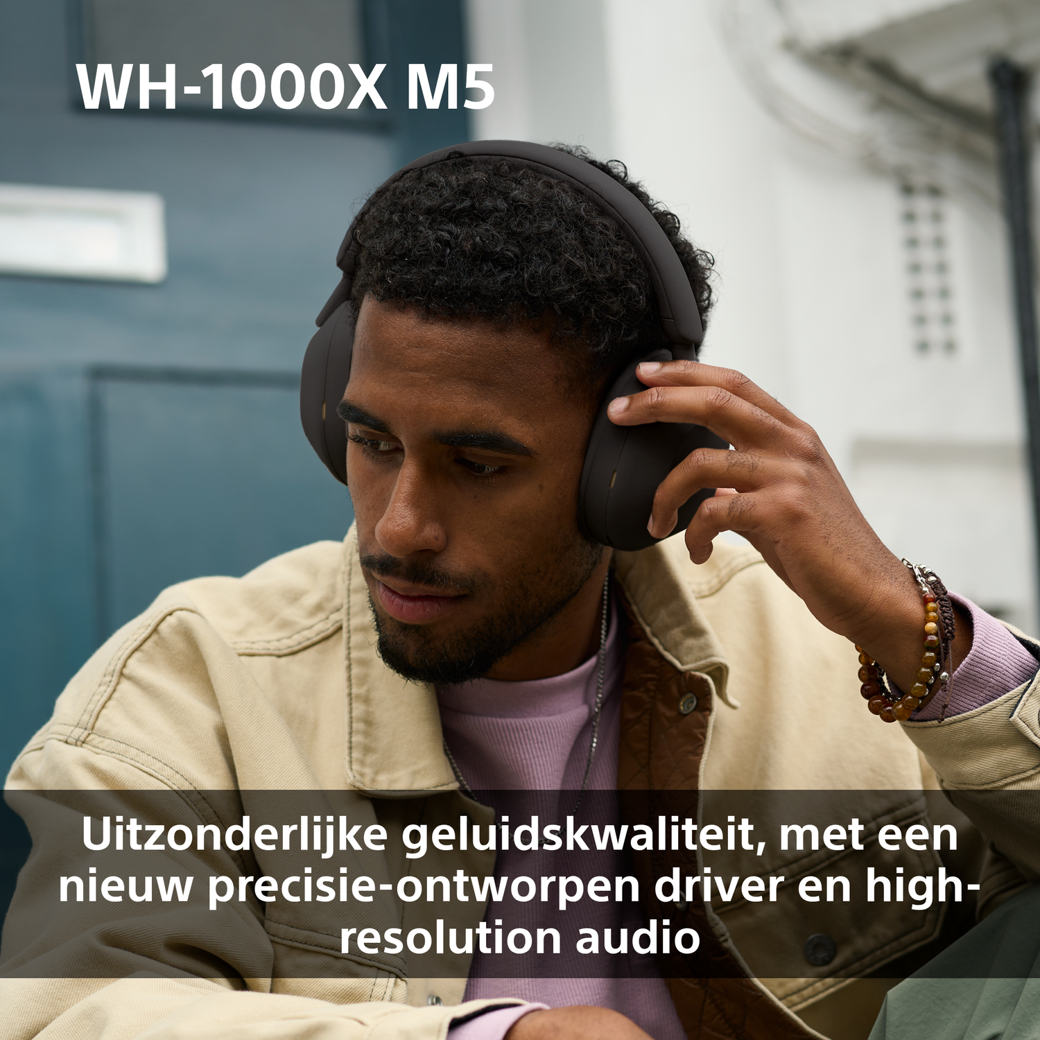 A man wearing black headphones holds one ear cup. He wears a tan jacket over a pink shirt. Text reads 'WH-1000X M5'.