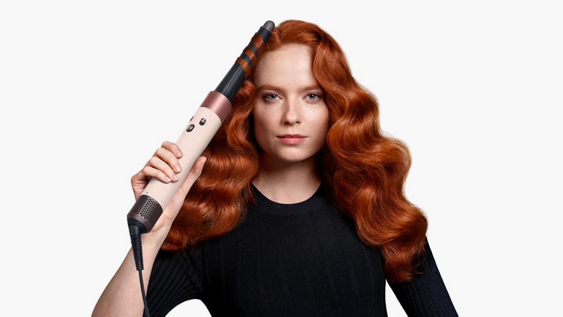 Une femme aux cheveux roux ondulés tient un fer à friser. Elle porte un haut noir. Le fond est blanc.