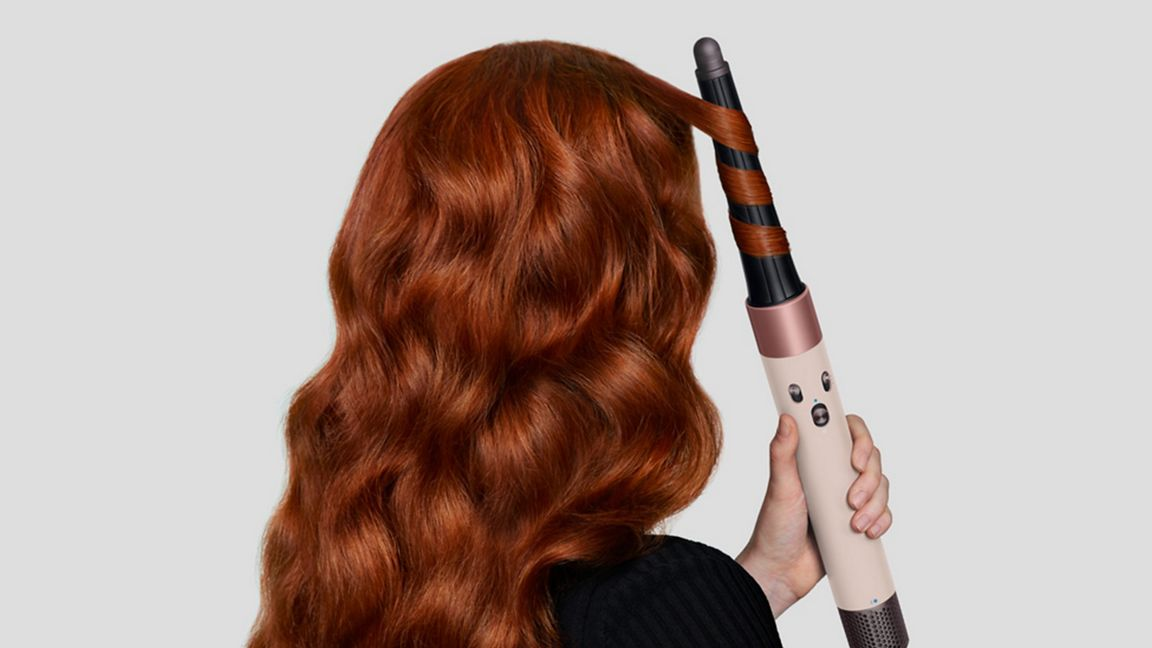 Une personne boucle des cheveux roux avec un fer à friser. Le fer est tenu dans la main droite. Fond blanc.