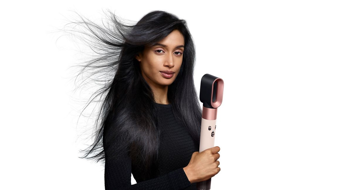 Une femme tient un sèche-cheveux. Ses cheveux noirs sont coiffés. Le sèche-cheveux est noir et or rose. Elle porte un haut noir.