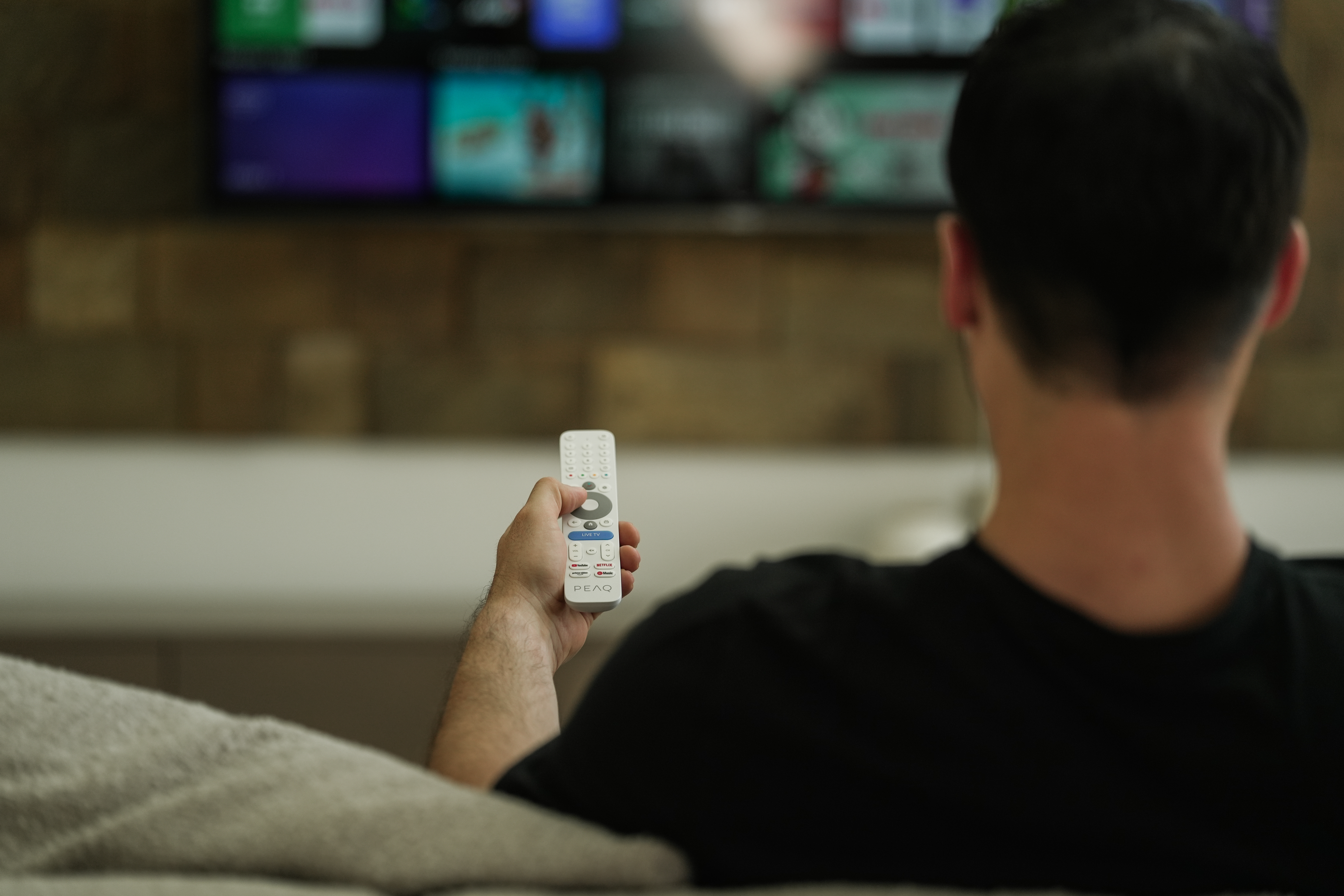 Eine Person hält eine Fernbedienung und schaut fern. Die Fernbedienung ist weiß. Der Fernseher ist an und zeigt verschiedene Apps. Die Person trägt Schwarz.