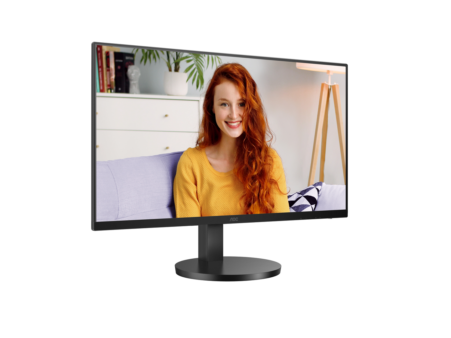 Una mujer con cabello rojo y un suéter amarillo sonríe en un monitor. El monitor está sobre un soporte negro. El fondo incluye muebles.