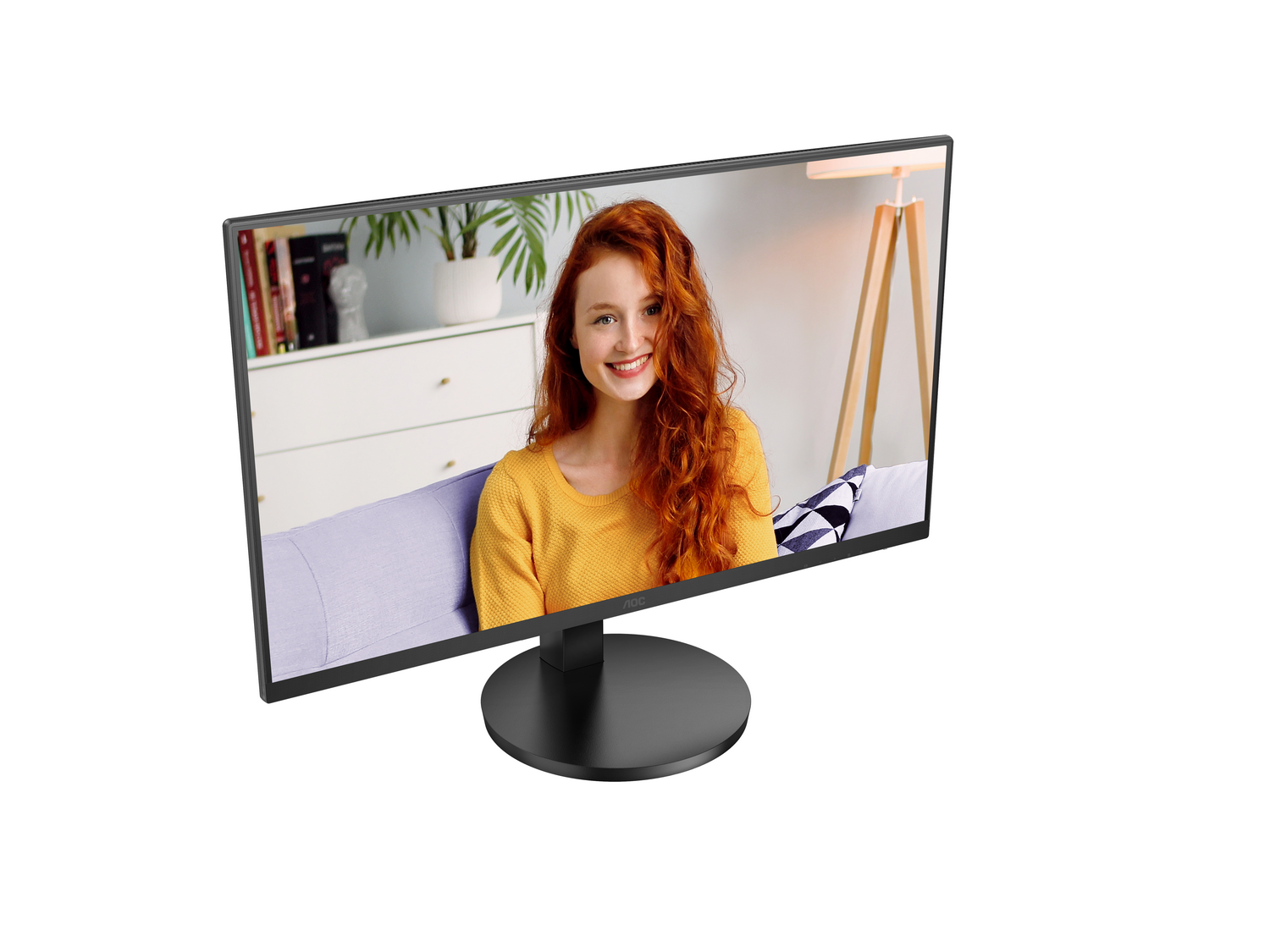 Un monitor de computadora muestra a una mujer pelirroja, con un suéter amarillo, sonriendo. Detrás hay una habitación con un sofá y una planta.