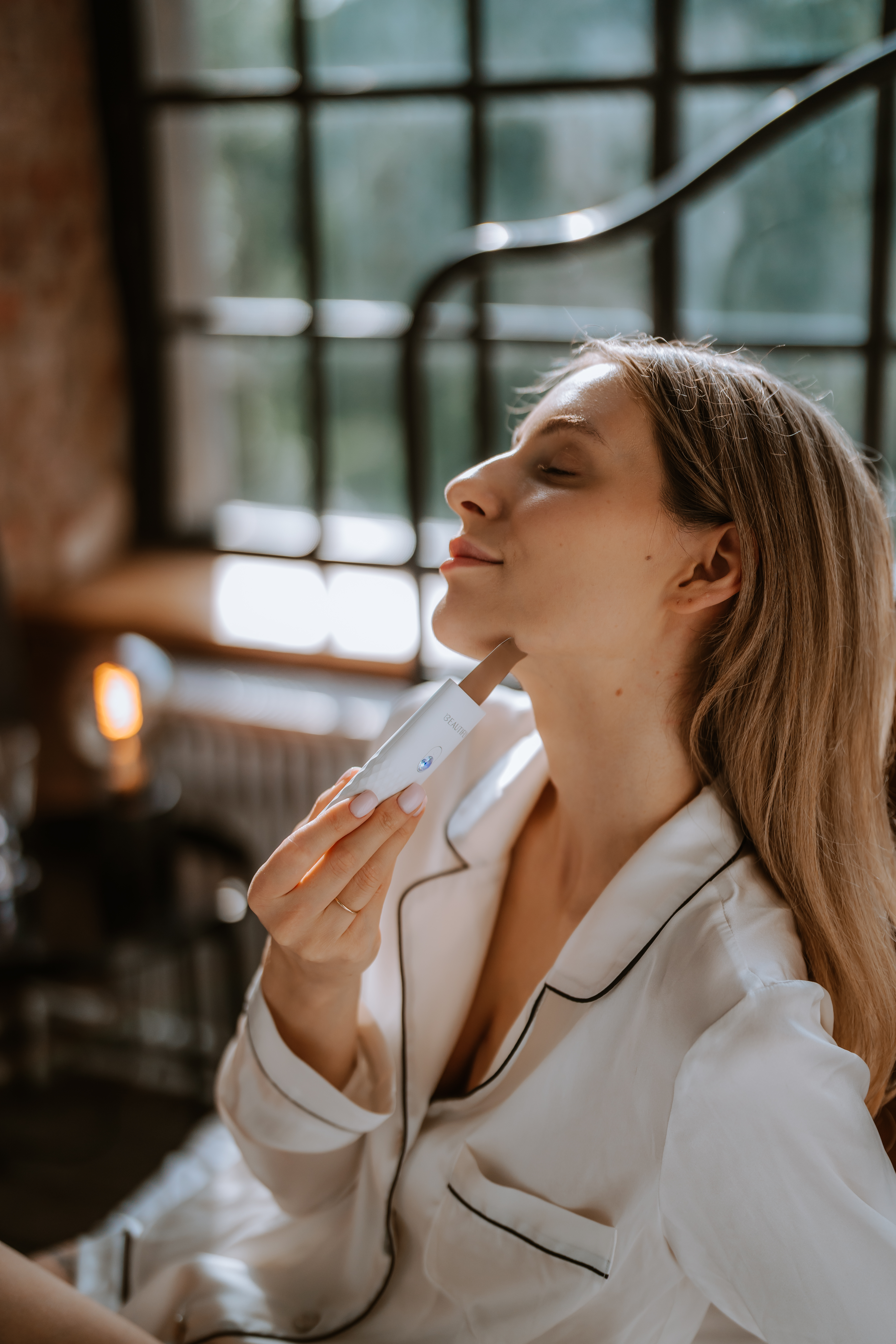 Eine Frau im weißen Bademantel verwendet ein Schönheitsgerät an ihrem Hals. Sie hat blondes Haar und einen sanften Ausdruck. Der Hintergrund ist unscharf.
