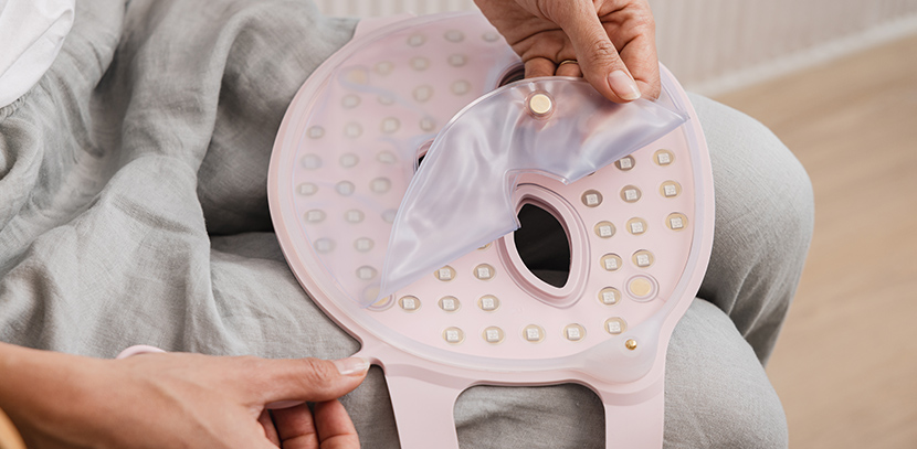 Eine Person hält eine rosa LED-Gesichtsmaske und löst eine transparente Plastikabdeckung ab. Die Person sitzt und trägt eine graue Hose, ein weißes Hemd ist sichtbar.