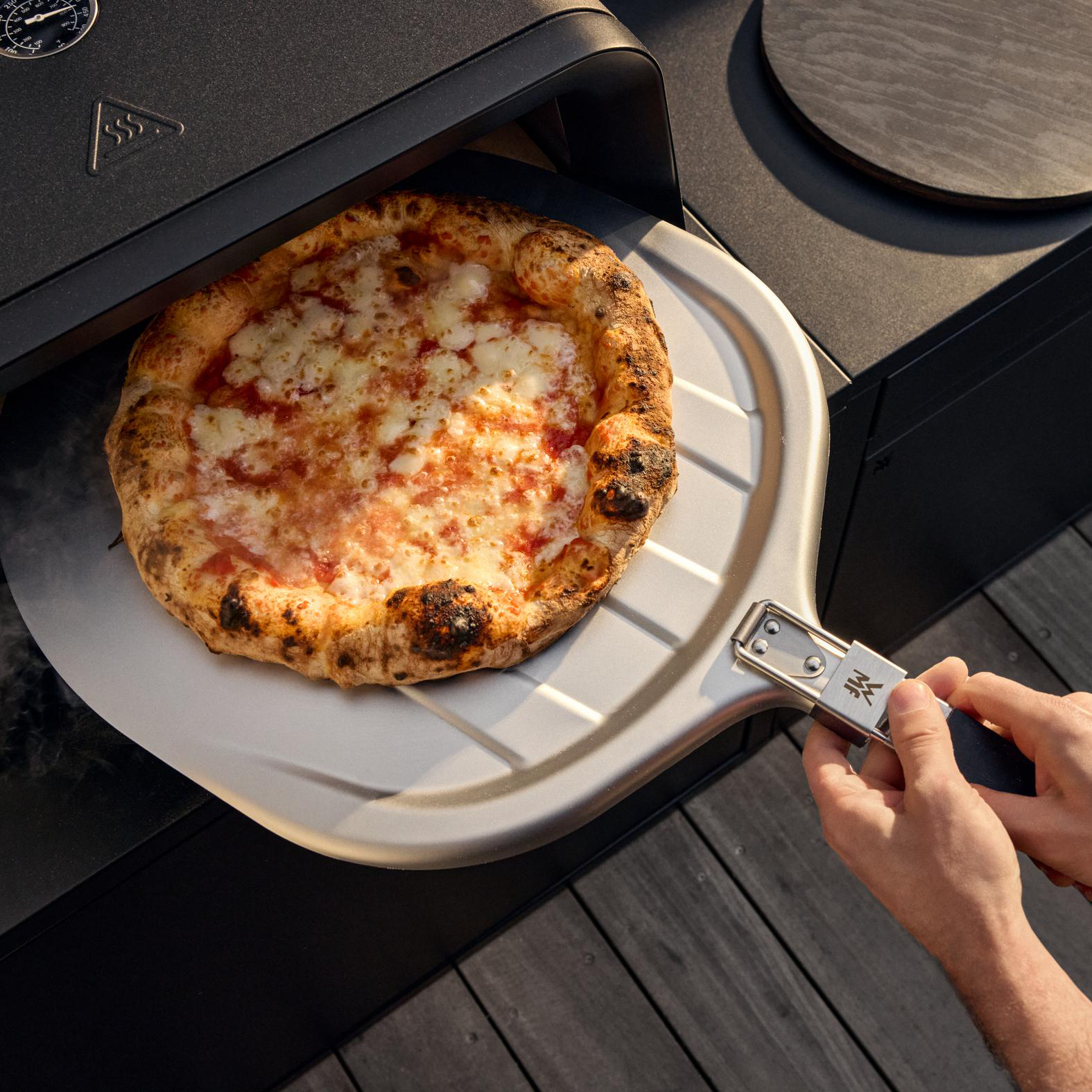 Eine Pizza mit Käse liegt auf einem Pizzaschieber. Hände halten den Schieber. Die Pizza befindet sich in der Nähe eines heißen Ofens.