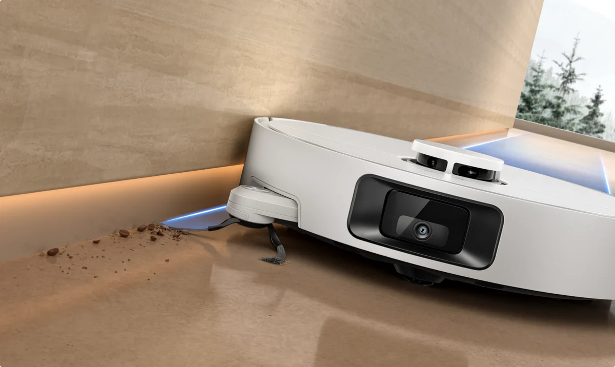 Egy fehér robotporszívó világosbarna felületet tisztít. Kávébabok és por a fal közelében. A háttérben egy ablak látható fákkal.