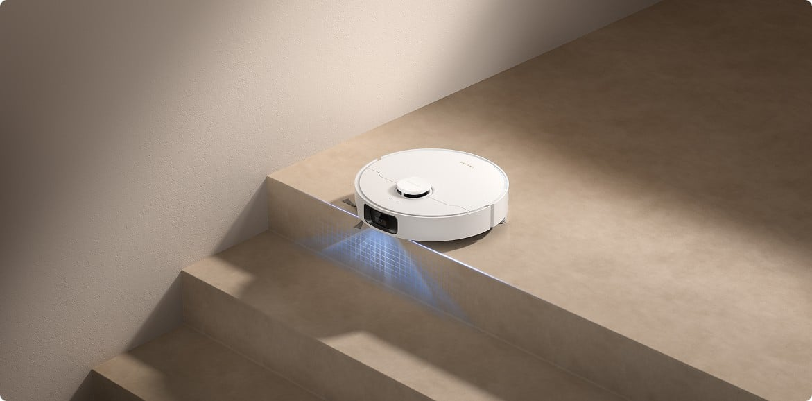 Egy fehér robotporszívó a lépcsőn. A padló és a falak bézsek. Kék fény világít a porszívóból.