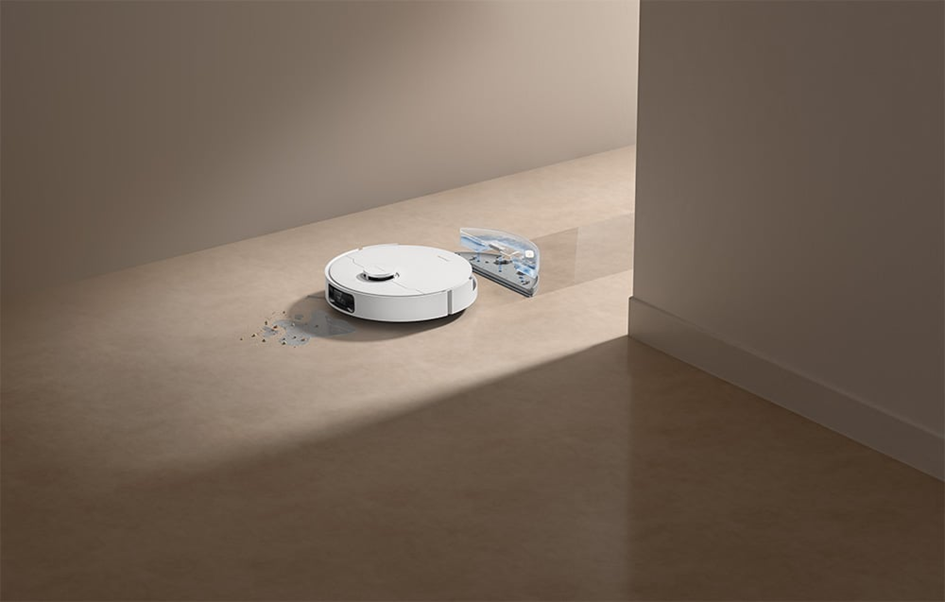 Egy fehér robotporszívó a padlón áll, közel egy átlátszó víztartályhoz. Por és törmelék van a porszívó körül. A jelenet beltéri.