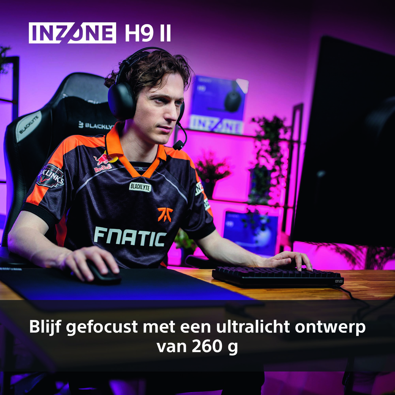 A man wearing a headset and gaming jersey sits at a desk with a keyboard and mouse. The background has a large screen and plants.