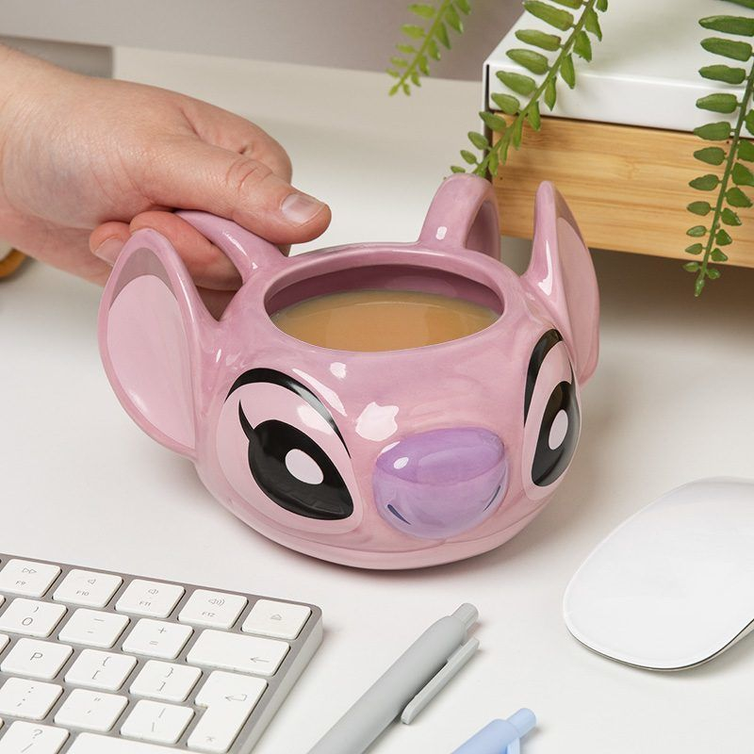 A pink Stitch mug with coffee sits on a white desk. A hand holds the mug. A keyboard, mouse, and pens are also on the desk.