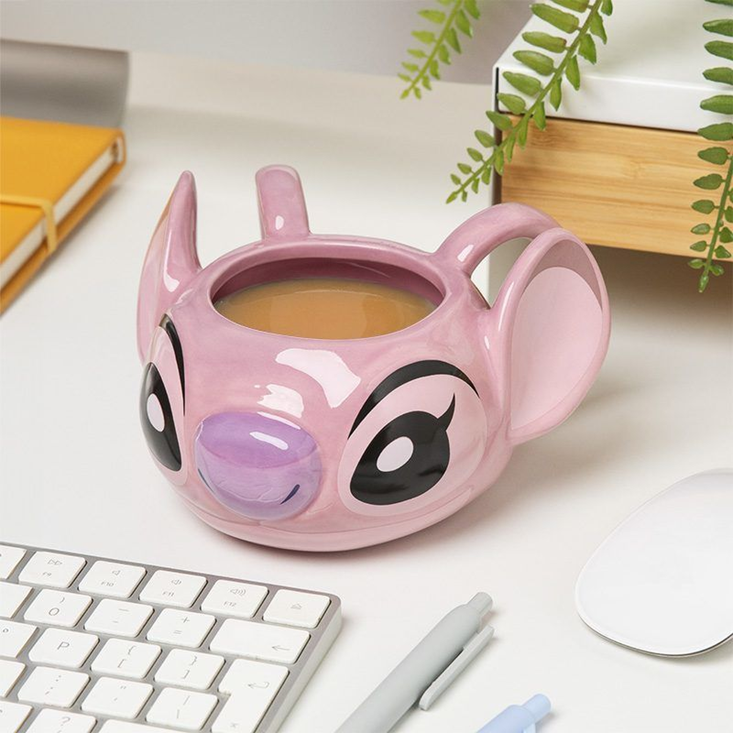 A pink Stitch mug filled with coffee sits on a white desk. A keyboard, pens, and mouse are nearby. Green plants are in the background.