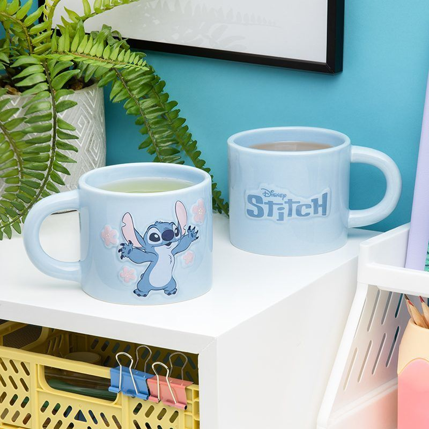 Two light blue mugs with Stitch design and filled with liquid, on a white shelf. A plant is in the background.