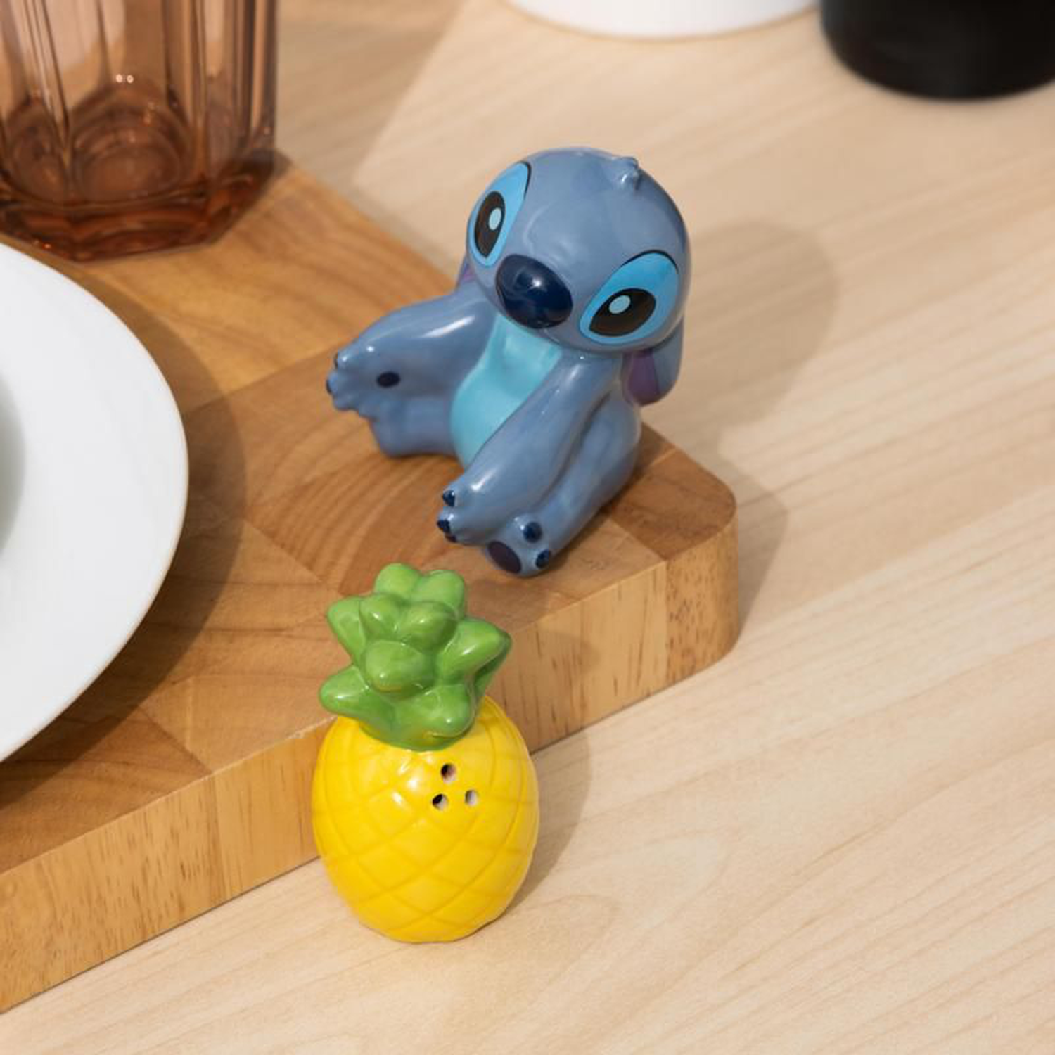 A Stitch figurine and a pineapple-shaped salt shaker rest on a wooden surface. A white plate and a glass are partially visible.