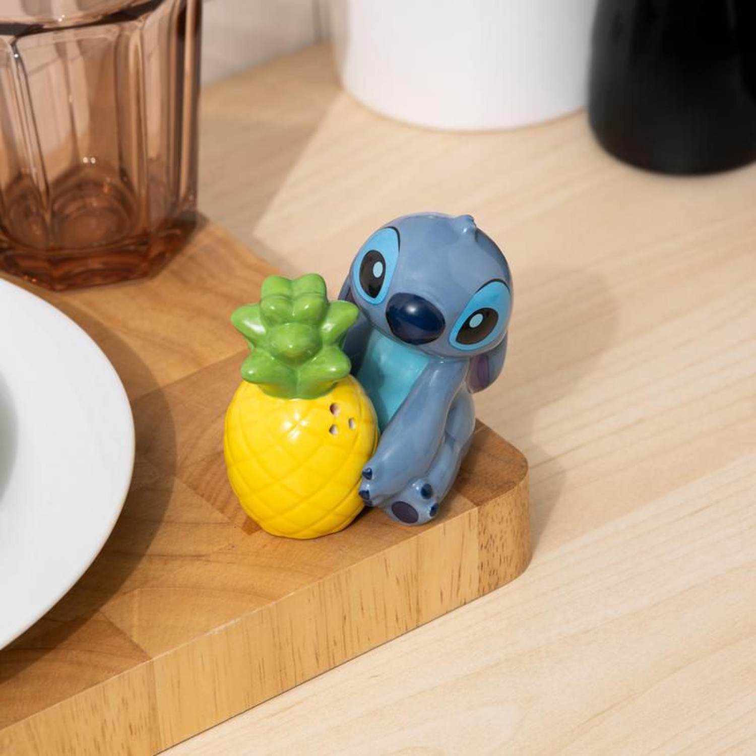 Stitch figure leaning against a pineapple-shaped salt shaker. Both are on a wooden board. A glass, plate and other objects are in the background.