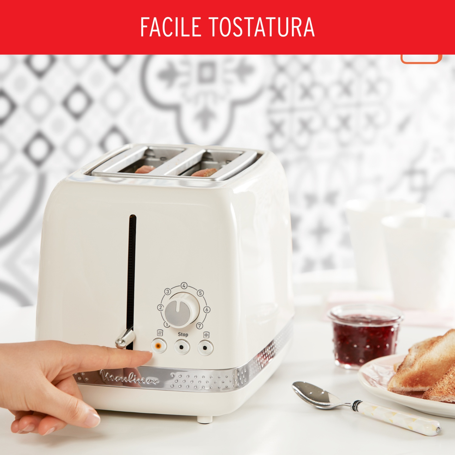 Un tostapane color crema con una mano che preme un pulsante. Toast, marmellata e un cucchiaio sono vicini. Lo sfondo ha un muro a motivi.