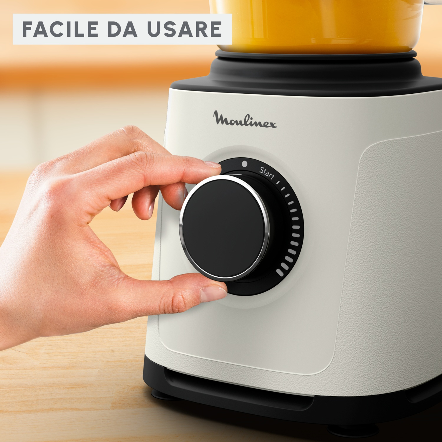 Una mano gira la manopola di un frullatore Moulinex bianco e nero. La manopola è nera con 'Start' sopra. Il frullatore contiene liquido arancione.