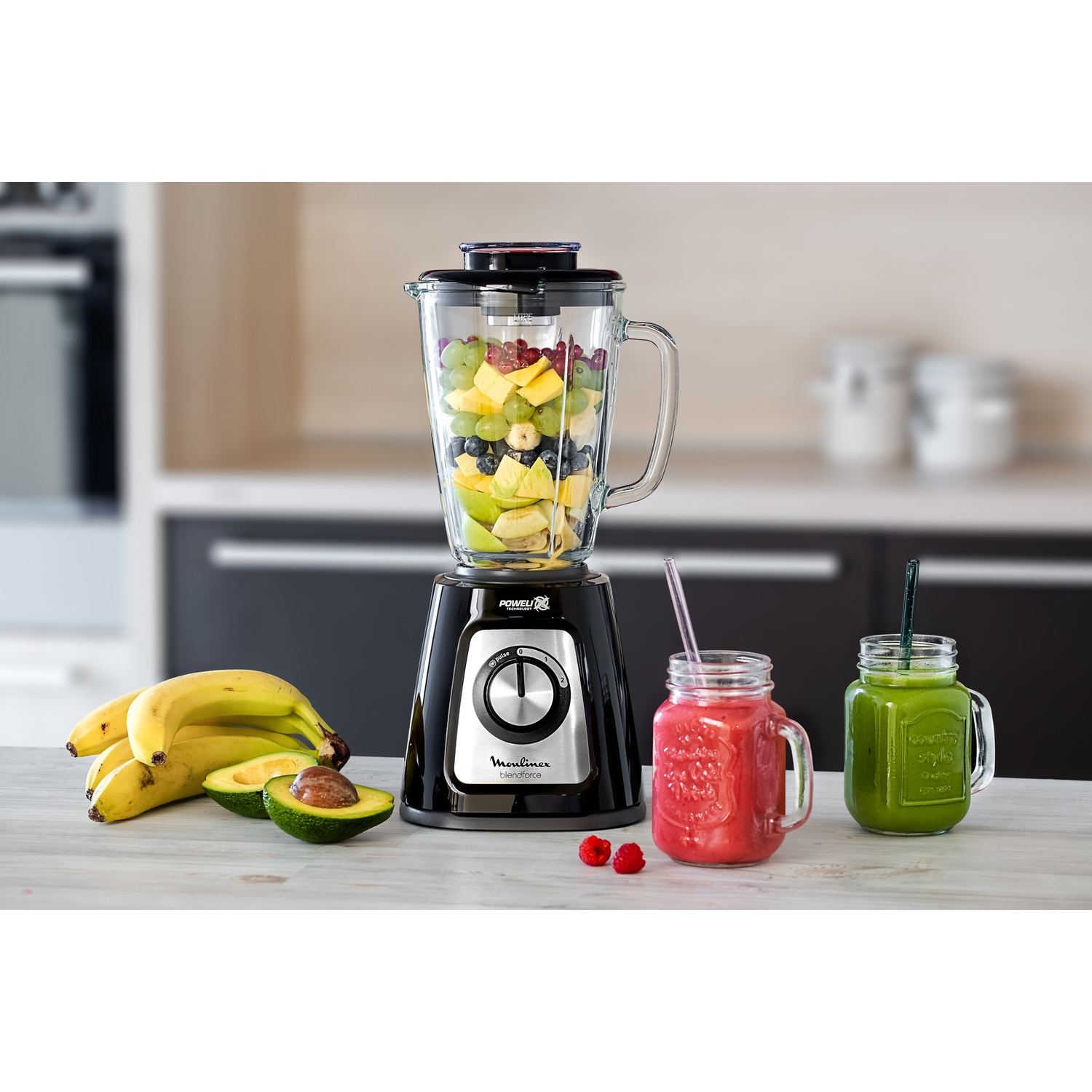 Un frullatore con frutta all'interno, accanto a bicchieri di frullato e frutta come banane e avocado.