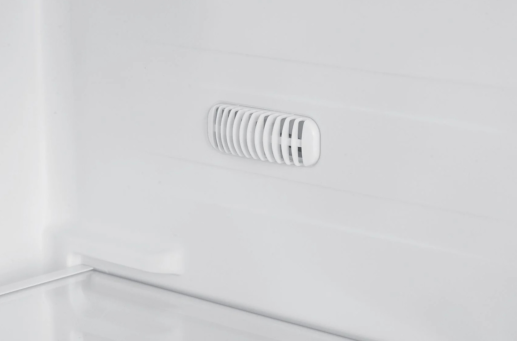 Wewnątrz białej lodówki widoczny jest otwór wentylacyjny z pionowymi listwami. Tłem jest białe wnętrze lodówki.