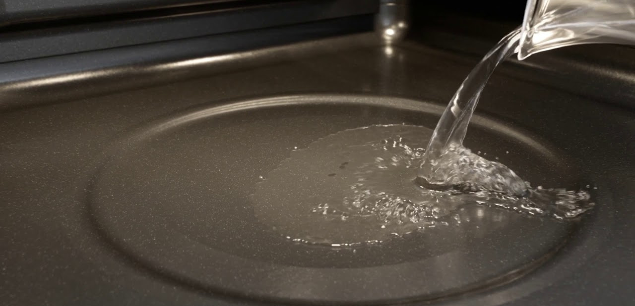 Woda wlewana do ciemnej patelni. Woda jest czysta, patelnia ma metaliczne wykończenie.