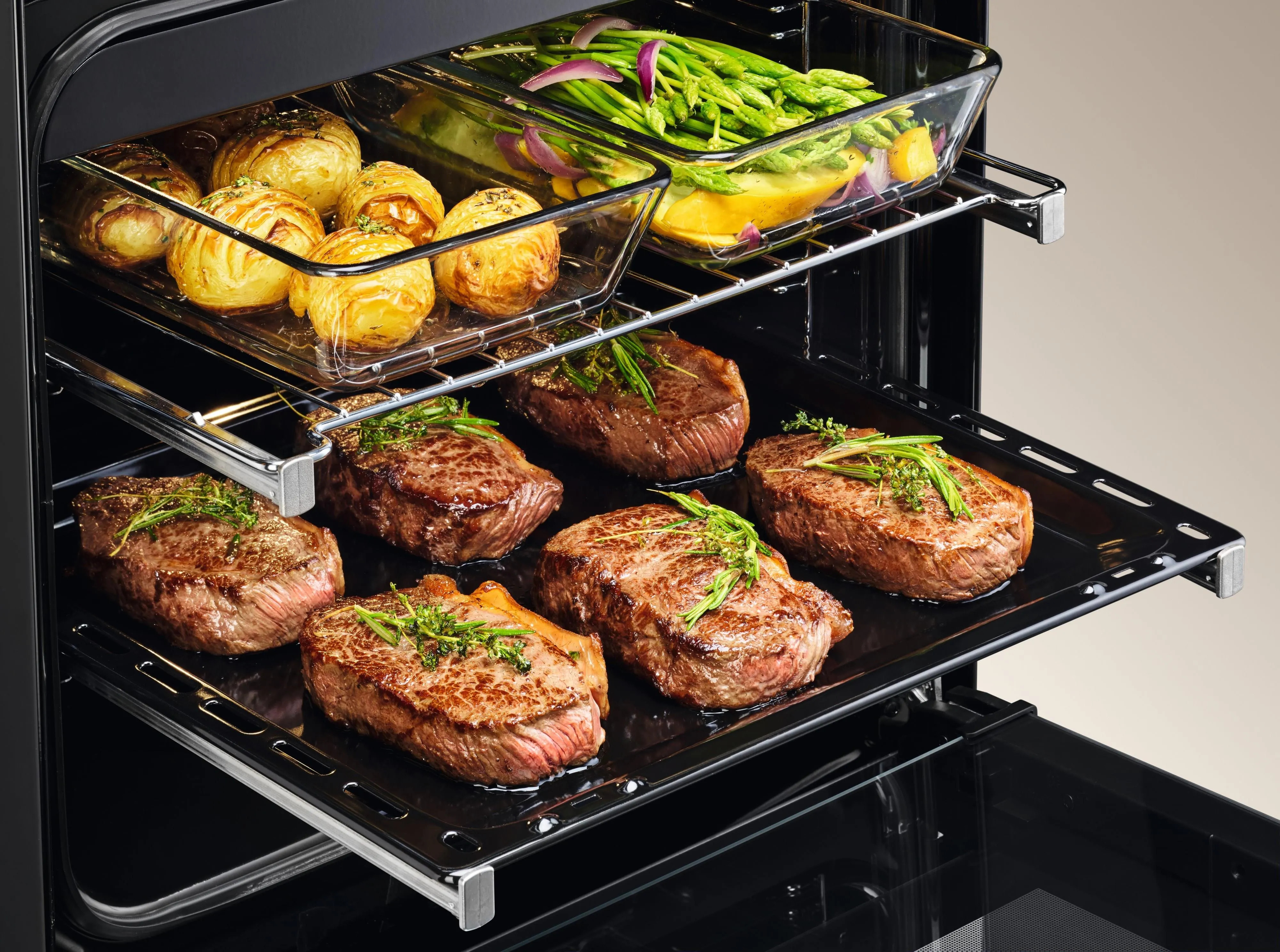 Piekarnik z ugotowanymi stekami i warzywami. Ziemniaki i szparagi są w szklanych naczyniach. Piekarnik jest czarny.