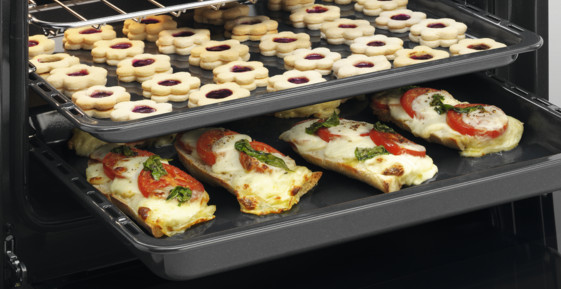 Piekarnik z dwiema tacami. Górna taca ma ciasteczka, dolna chleb z pomidorami i serem.