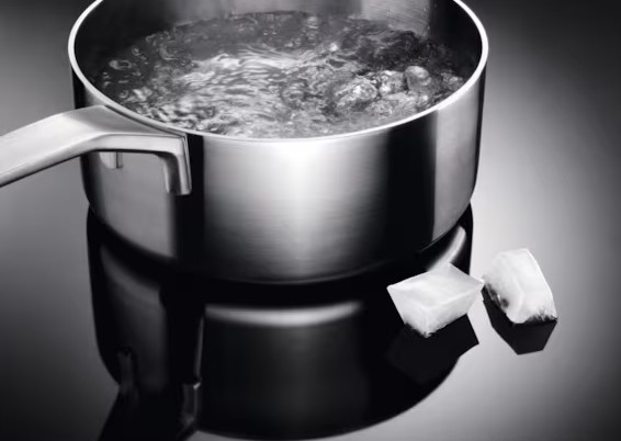 Garnek ze stali nierdzewnej z wrzącą wodą i dwiema kostkami lodu na odblaskowej powierzchni. Garnek ma uchwyt.