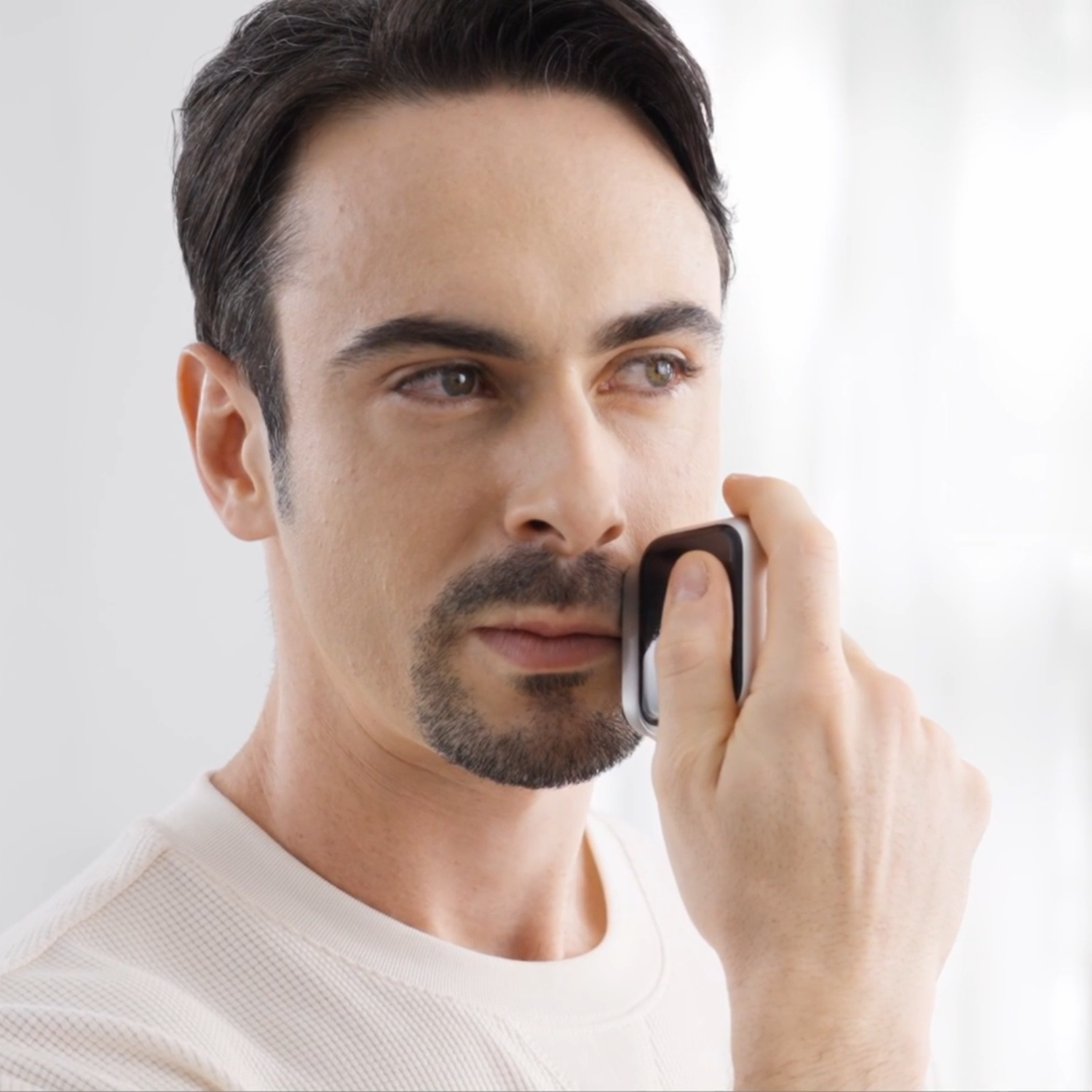 A man shaves his face with a black and silver electric shaver. He is wearing a white shirt. The background is blurred.