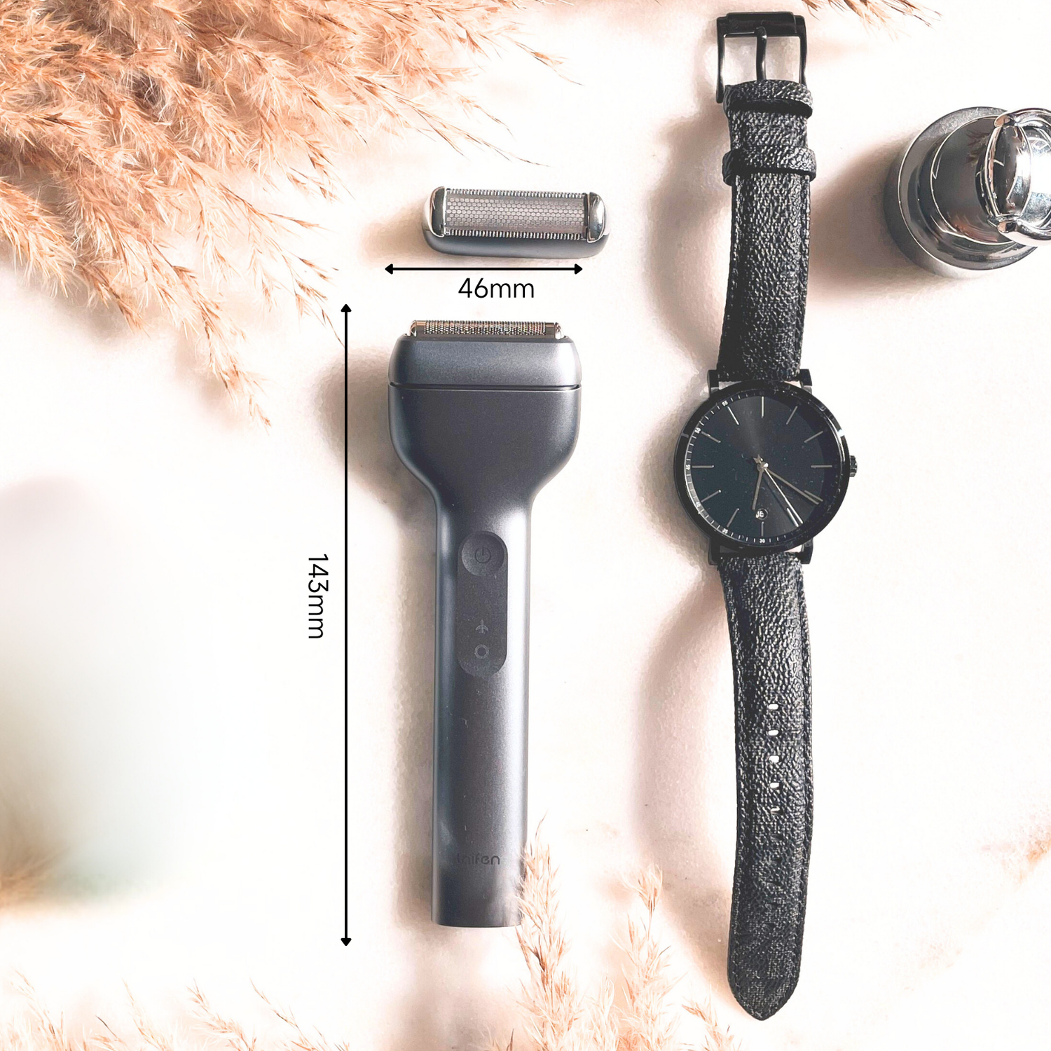 Un rasoir électrique, une tête de rasoir, une montre-bracelet noire et un petit objet chromé sur une surface blanche.