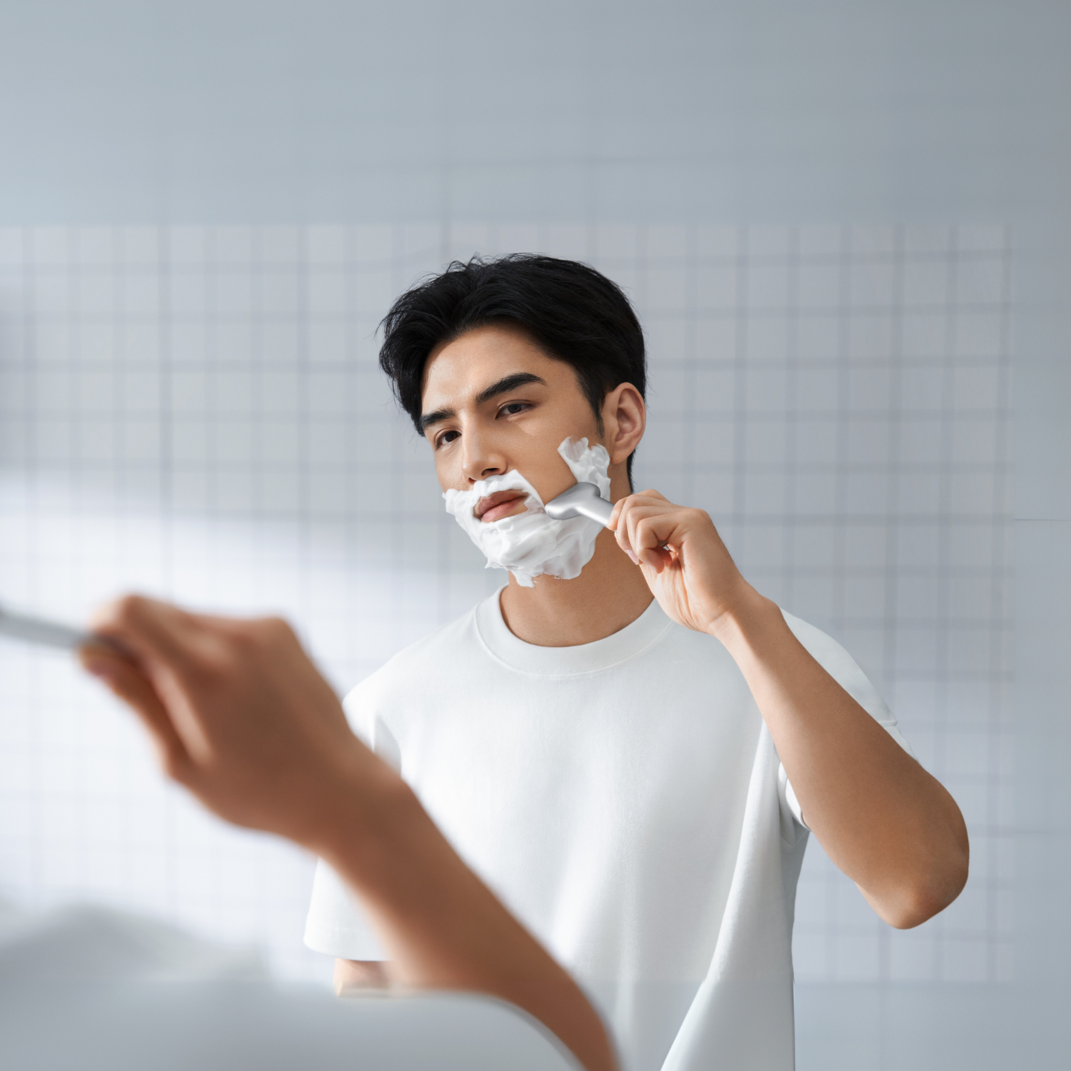 A man shaves his face in a bathroom. He is wearing a white t-shirt and using a razor. His face is covered in shaving cream.